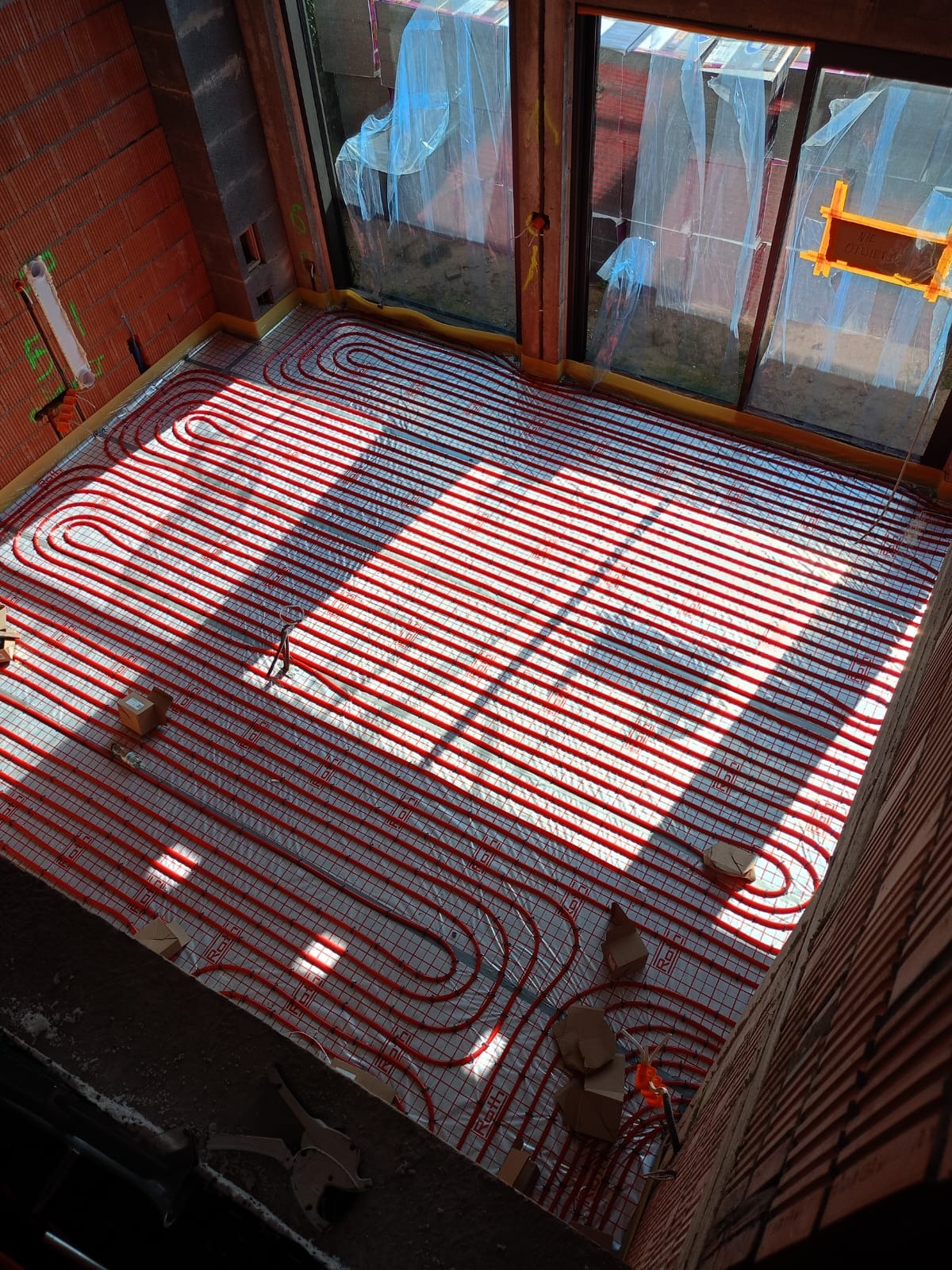 Czerwone rurki systemu grzewczego ułożone na siatce w surowym pomieszczeniu z ceglanymi ścianami, widoczne okna z folią ochronną i narzędzia pracy.