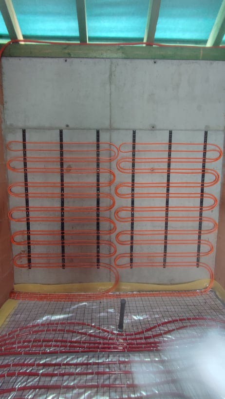 Montaż ogrzewania podłogowego: pomarańczowe rurki rozprowadzone na siatce izolacyjnej z widocznym pionowym elementem instalacji, mocowane do ściany z betonu komórkowego za pomocą czarnych uchwytów.