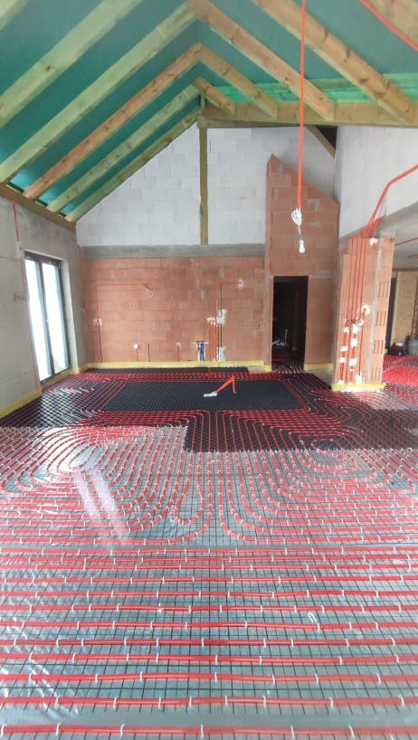 Montaż czerwonych rurek w systemie ogrzewania podłogowego na metalowej siatce w surowym wnętrzu z widoczną konstrukcją dachu i ścianami z cegły.