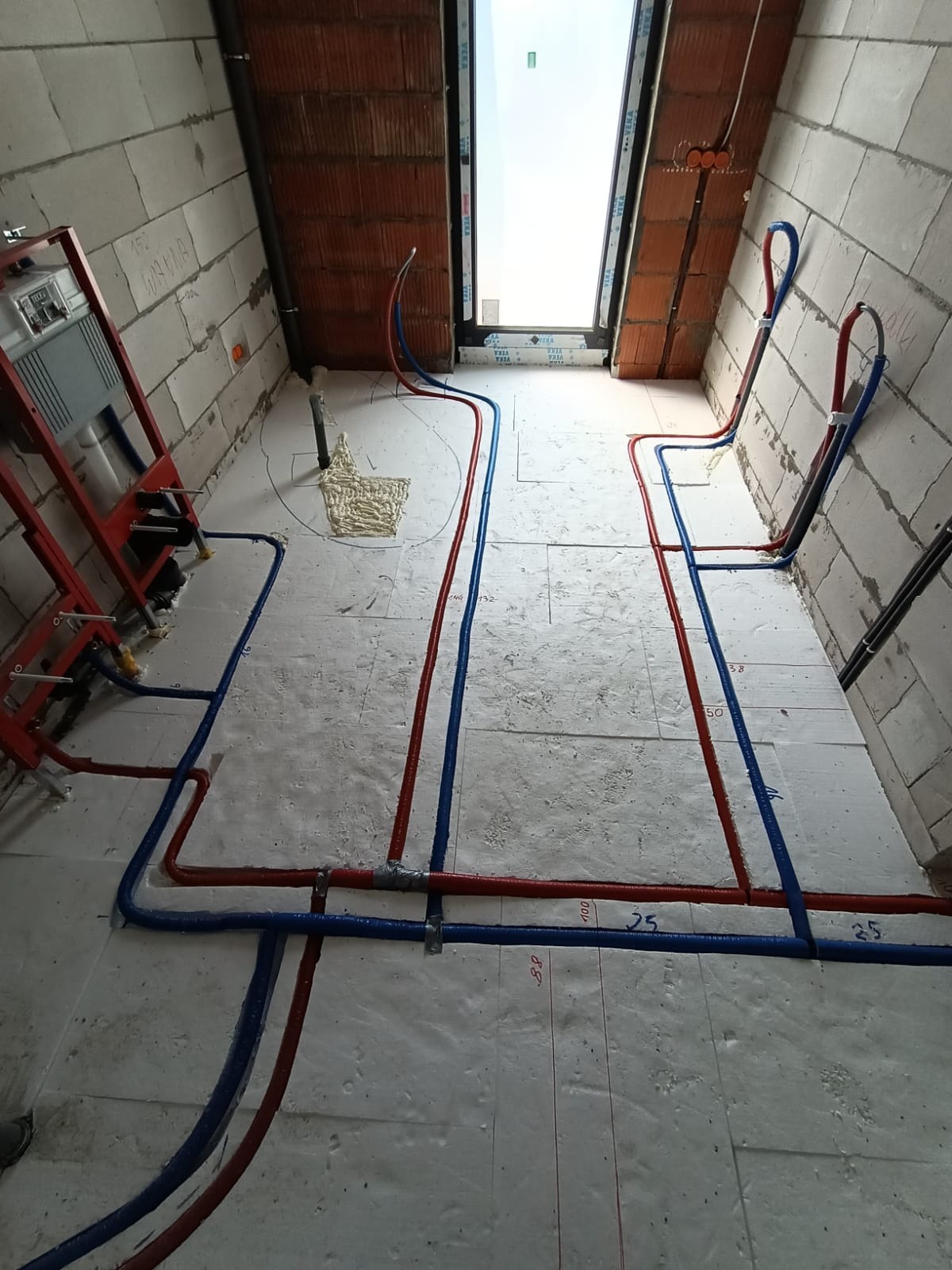 Wnętrze poddasza w trakcie montażu instalacji wodnej i grzewczej; widoczne czerwone i niebieskie rury prowadzone po białej izolacji podłogowej, mocowane uchwytami; w tle okno dachowe i ściany...