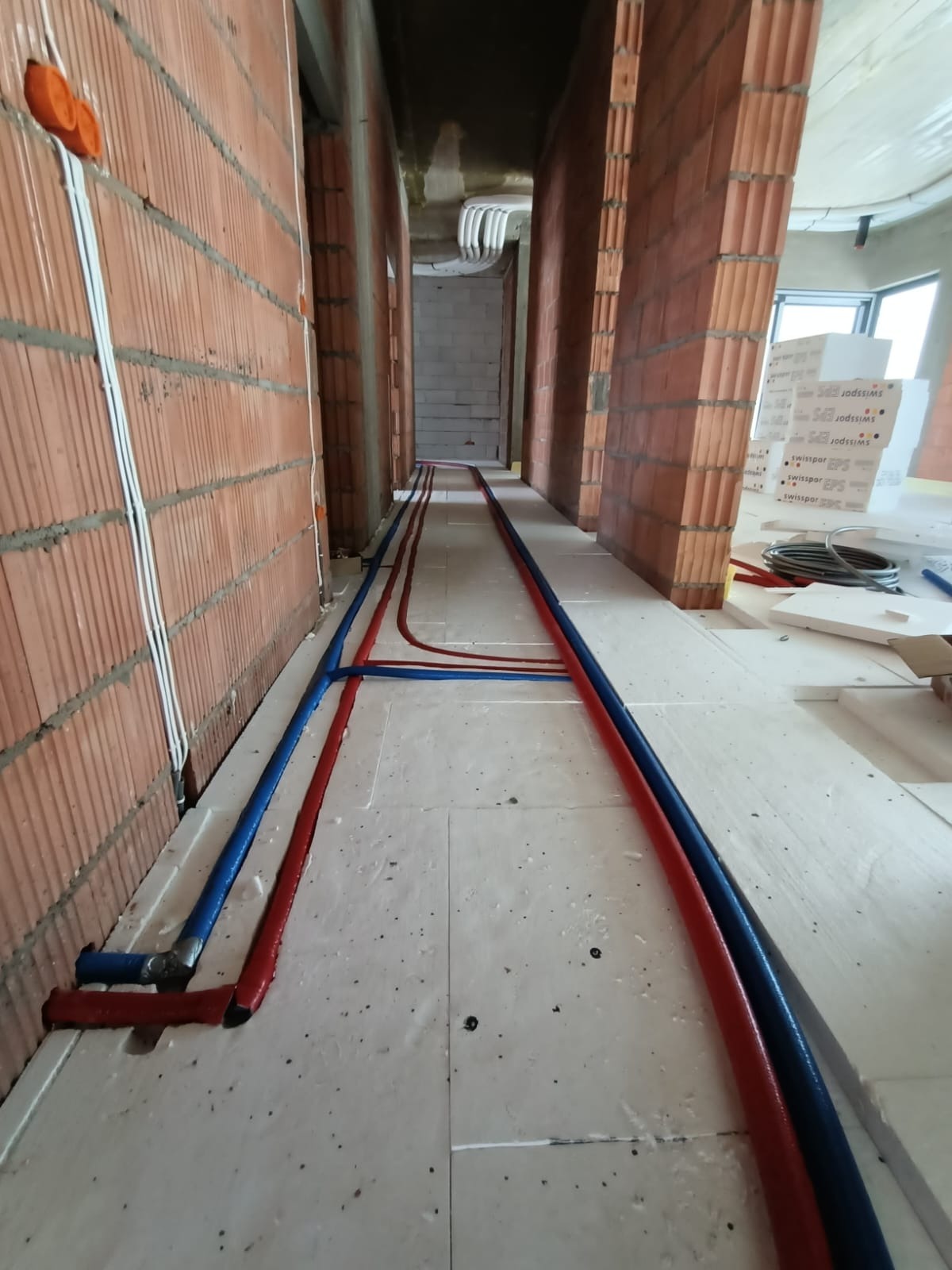 Widok korytarza w trakcie budowy, z widocznymi czerwonymi i niebieskimi rurami ułożonymi na białych płytach izolacyjnych, prowadzącymi wzdłuż ścian z czerwonej cegły.