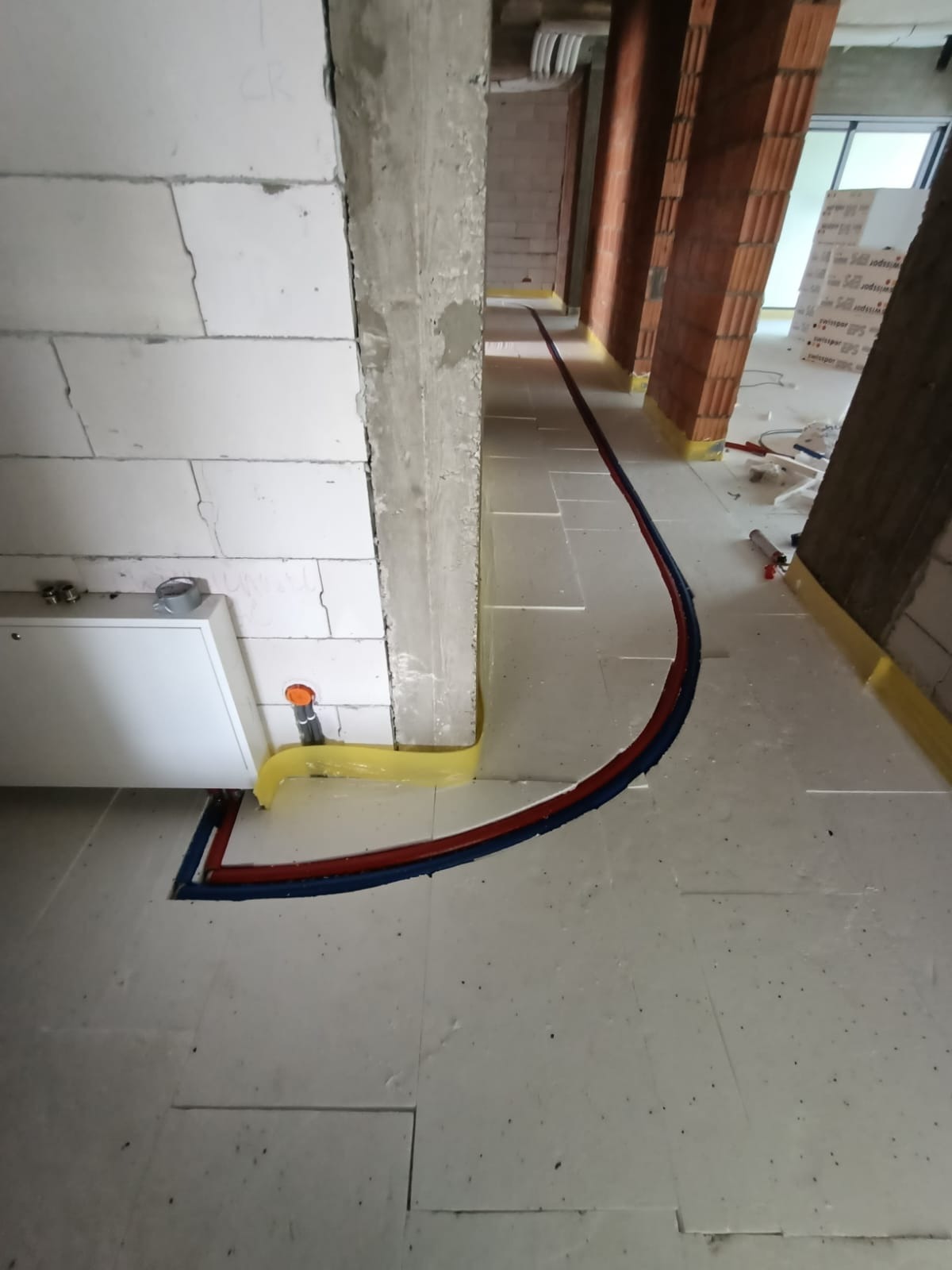 Instalacja rur grzewczych w izolacji podłogowej, widoczne rury czerwone i niebieskie, białe ściany z betonu komórkowego, żółta taśma zabezpieczająca.