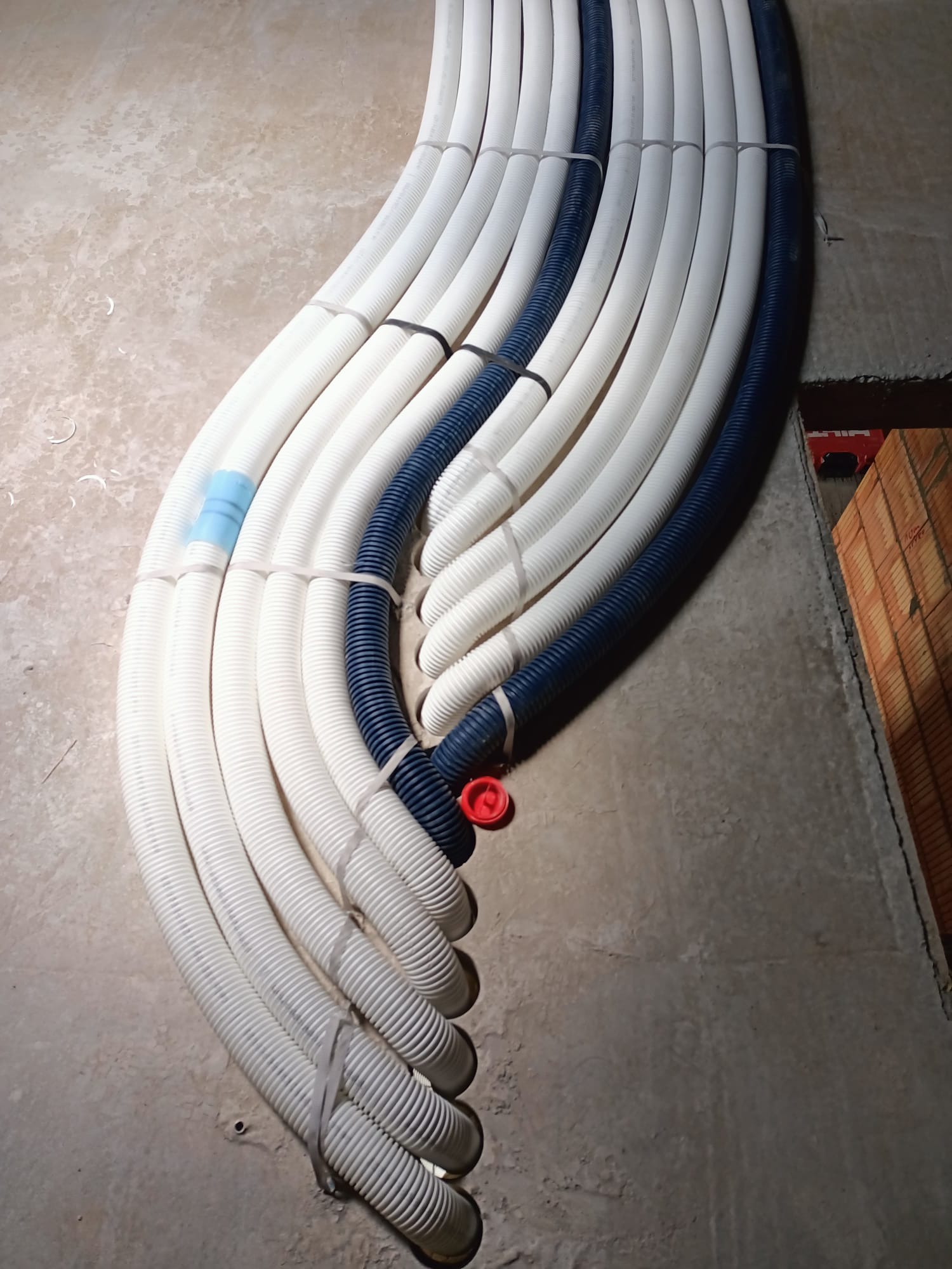 Rzędy białych i granatowych karbowanych rur, spiętych plastikowymi opaskami, ułożone na szarej, surowej podłodze w trakcie instalacji.
