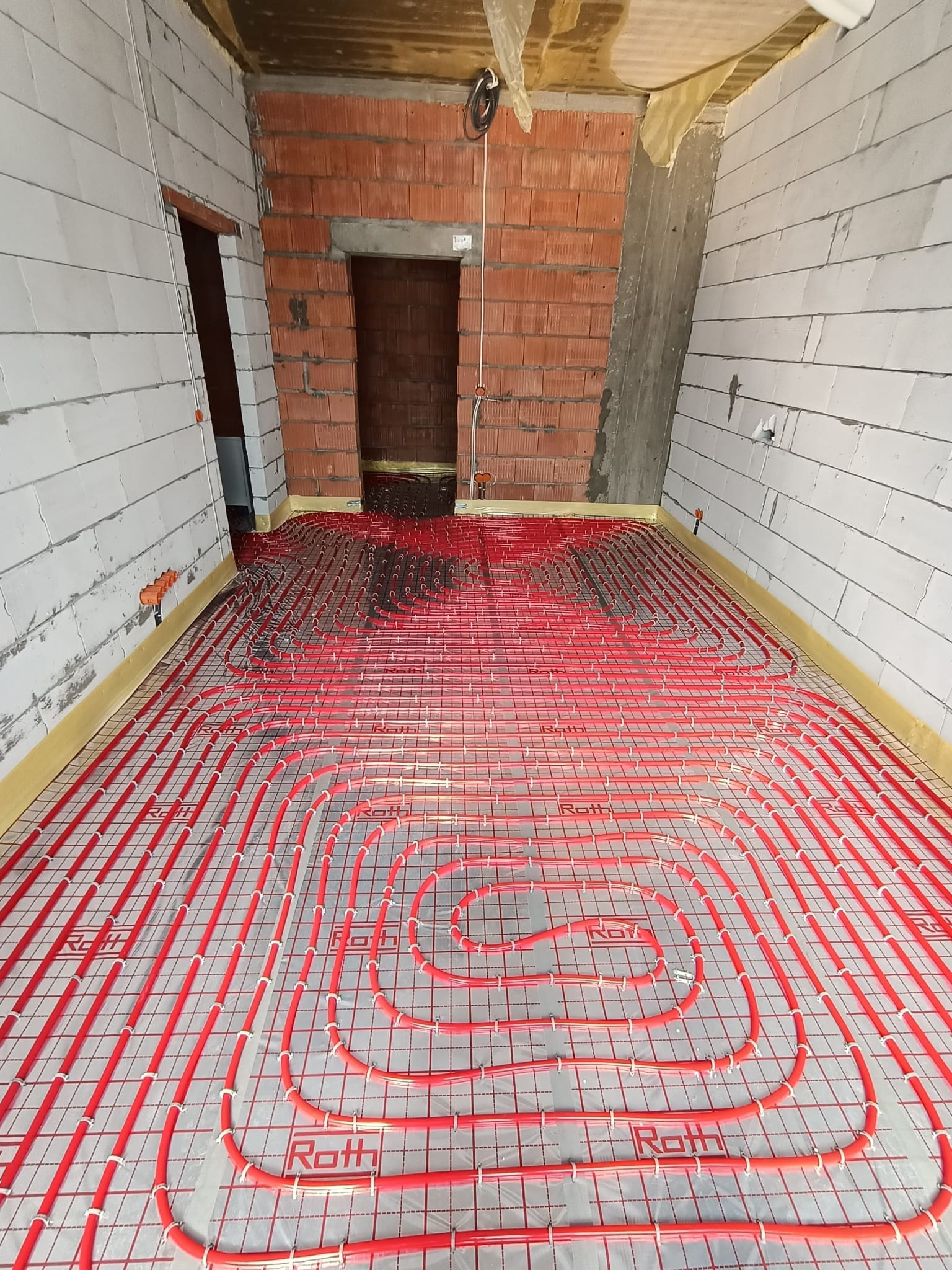 Czerwone rury systemu grzewczego ułożone spiralnie na podłodze, widoczne białe ściany z pustaków i fragment ściany z czerwonej cegły, w tle otwór drzwiowy.