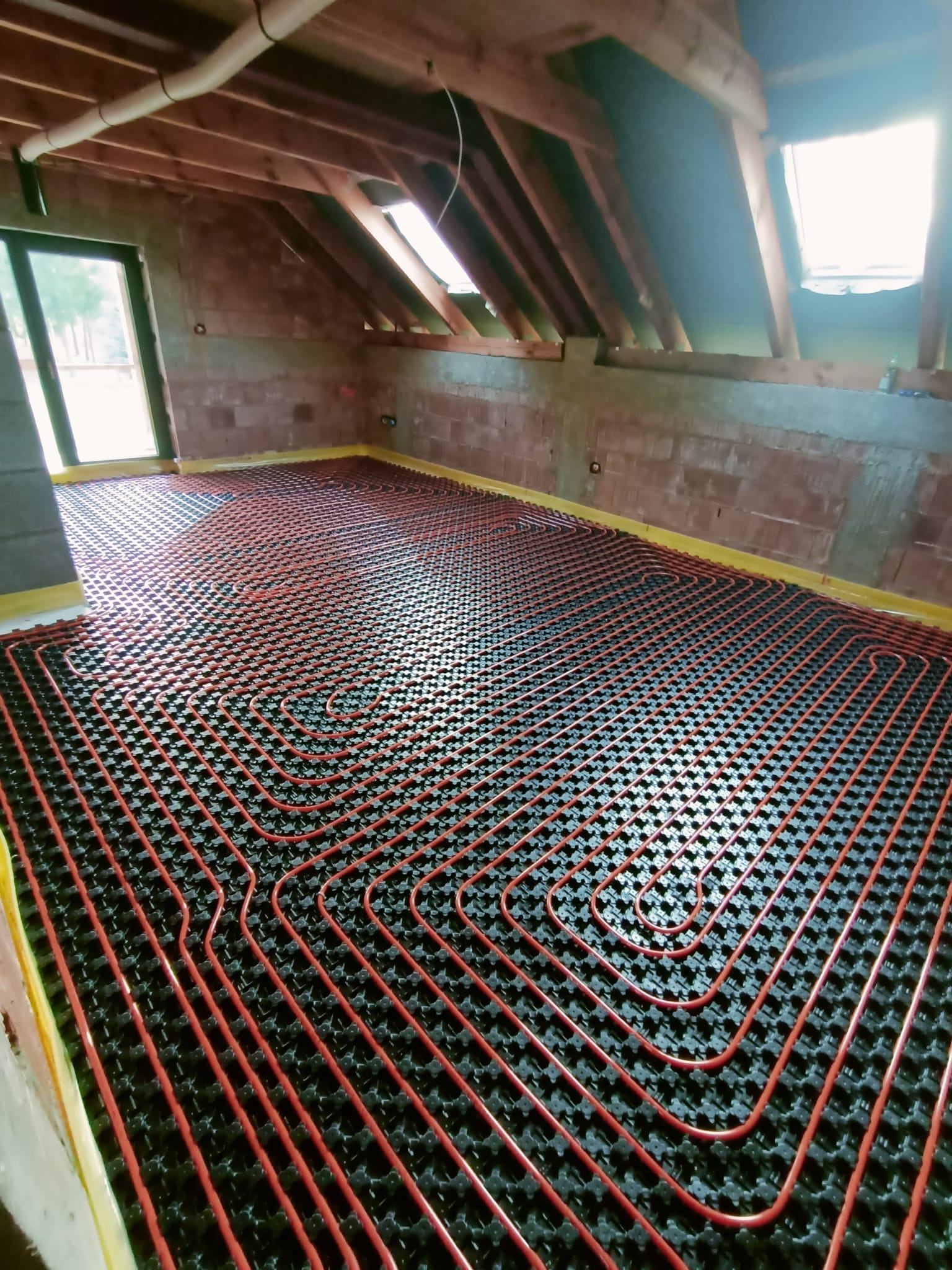 Instalacja ogrzewania podłogowego w surowym stanie, widoczne rury grzewcze ułożone na czarnej macie izolacyjnej, żółta taśma na krawędziach ścian, surowe ściany z pustaków i drewniana konstrukcja...