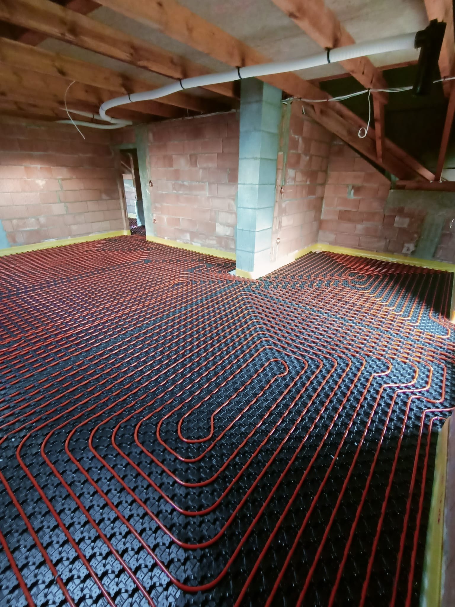 Czerwone rurki systemu rozprowadzenia ciepła ułożone na czarnej macie izolacyjnej w surowym wnętrzu z ceglanymi ścianami i drewnianym stropem.