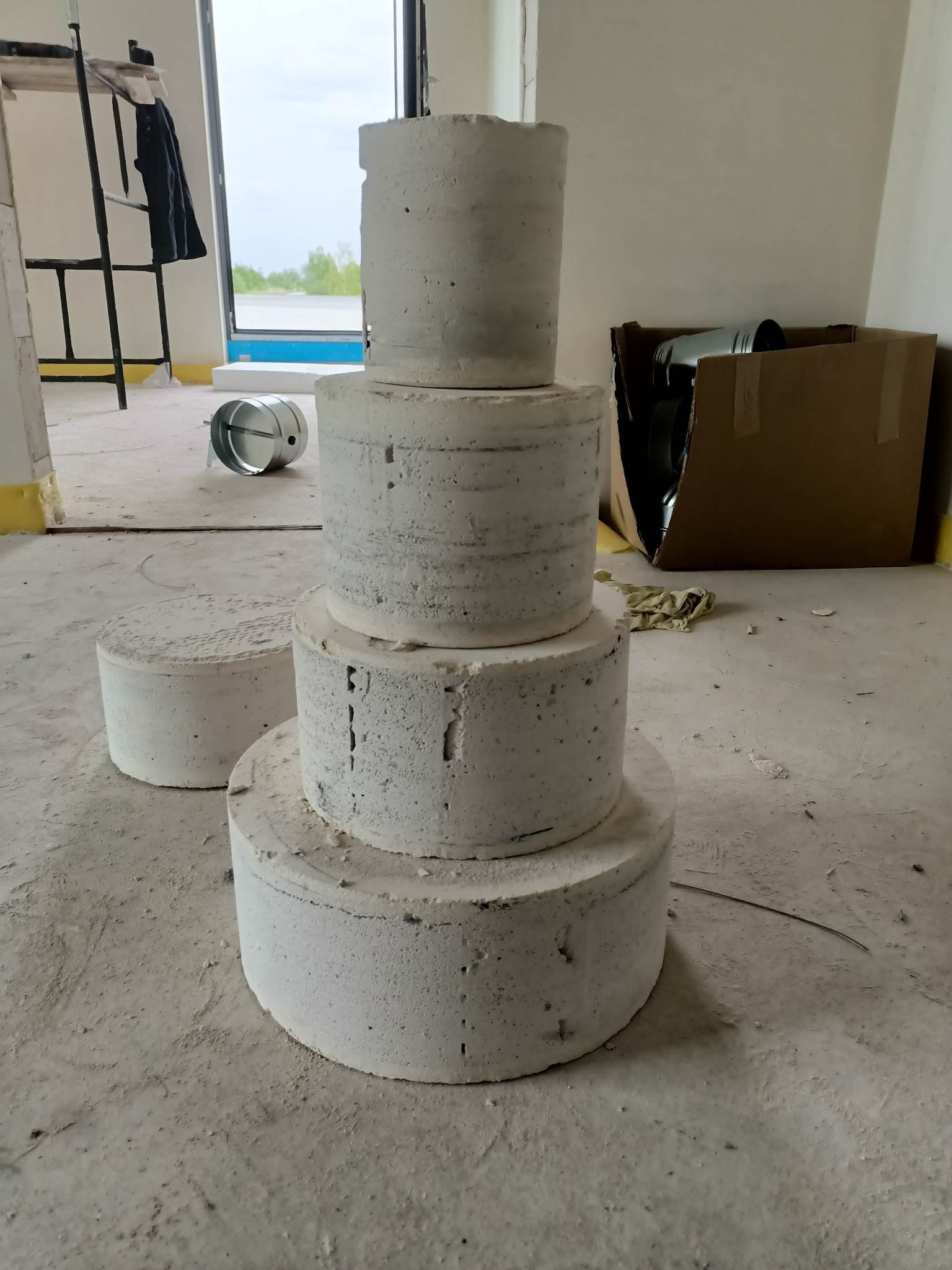 Cztery betonowe kręgi ułożone jeden na drugim w pomieszczeniu w trakcie remontu, widoczny metalowy kanał wentylacyjny i karton, w tle drabina i okno.
