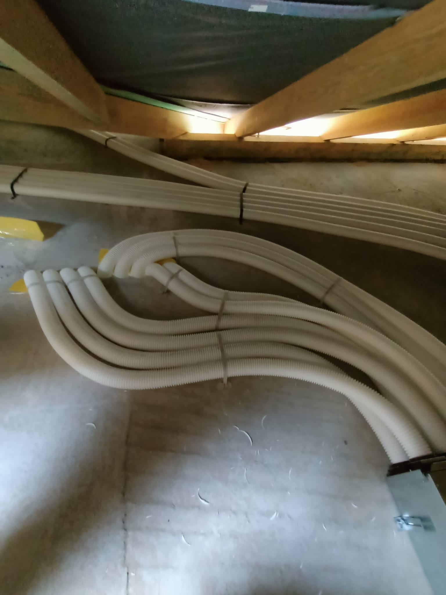 Białe, karbowane rury ułożone pod drewnianą konstrukcją stropu, prawdopodobnie w trakcie instalacji systemu rozprowadzającego ciepło.