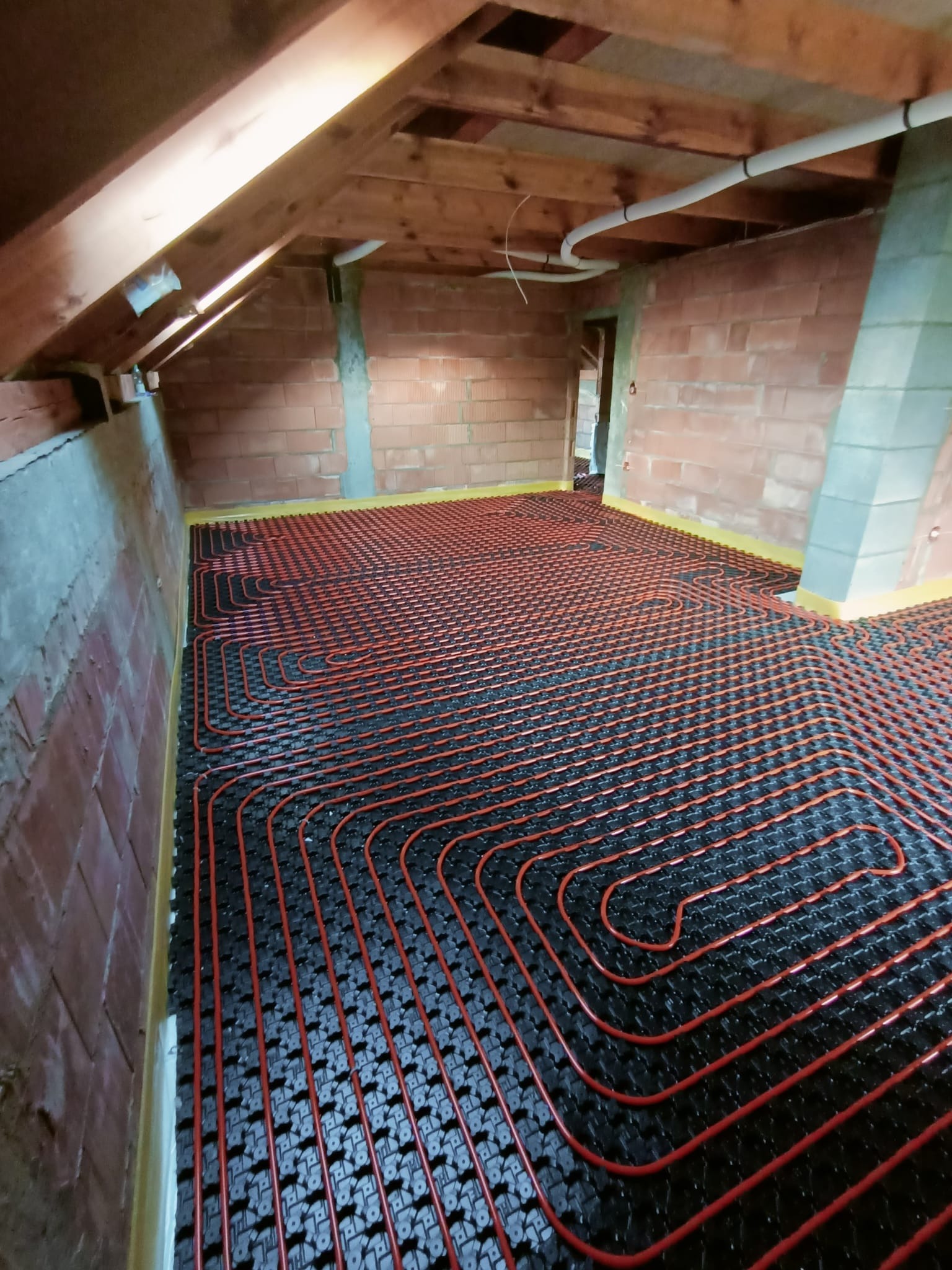 Instalacja ogrzewania podłogowego w surowym wnętrzu poddasza z widoczną czerwoną rurą grzewczą ułożoną na czarnej macie izolacyjnej, ceglane ściany i drewniana konstrukcja dachu.