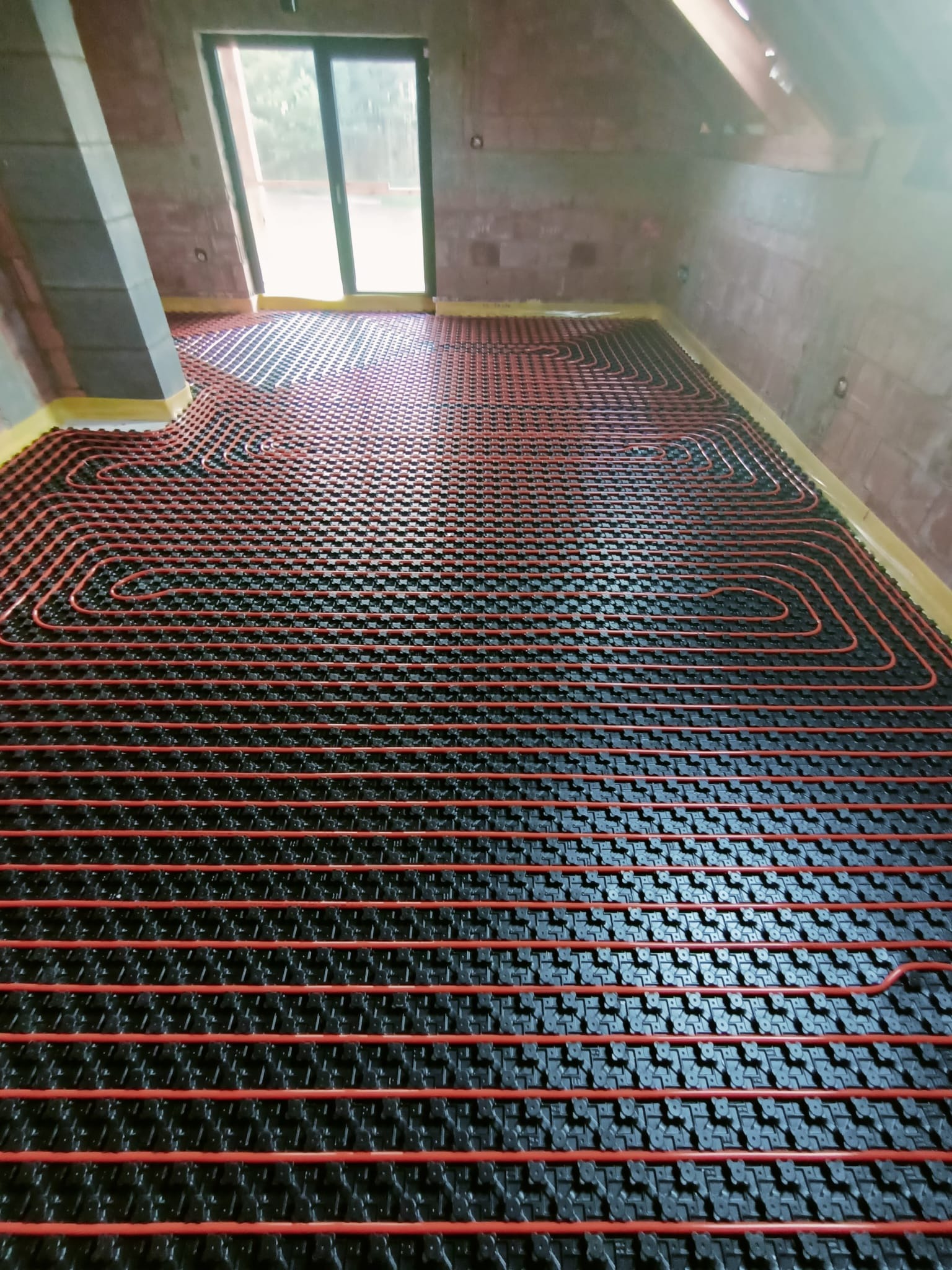 Czerwone rury układane w spiralny wzór na czarnej macie grzewczej, przygotowane do wylewki w surowym wnętrzu poddasza z oknem balkonowym.