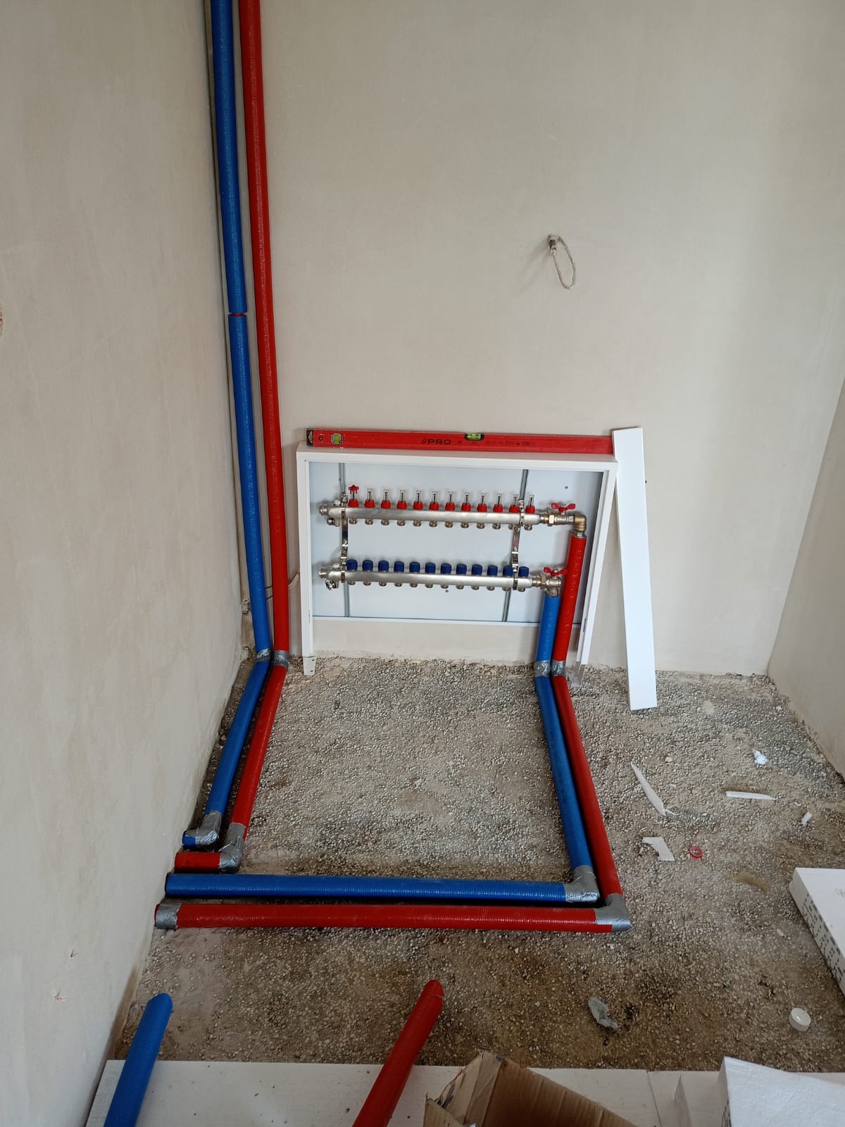 Rozdzielacz do instalacji centralnego ogrzewania podłogowego z rurami w kolorze czerwonym i niebieskim, zamontowany w surowym pomieszczeniu z podłożem wysypanym żwirem.