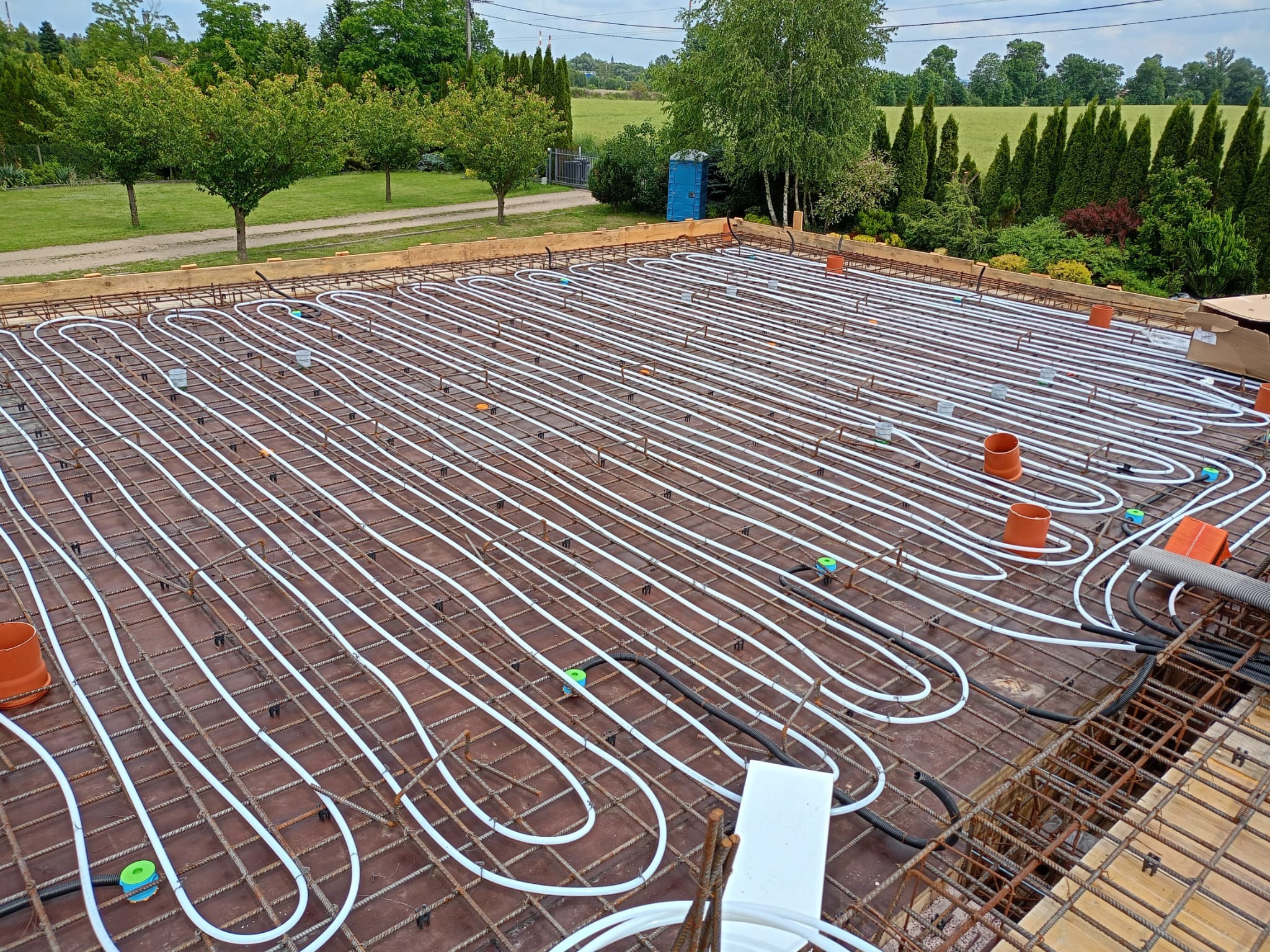 Białe rurki instalacji grzewczej ułożone w pętle na brązowej płycie z siatką zbrojeniową, widoczne pomarańczowe kształtki i elementy mocujące, w tle zielony krajobraz z drzewami i budynkami.
