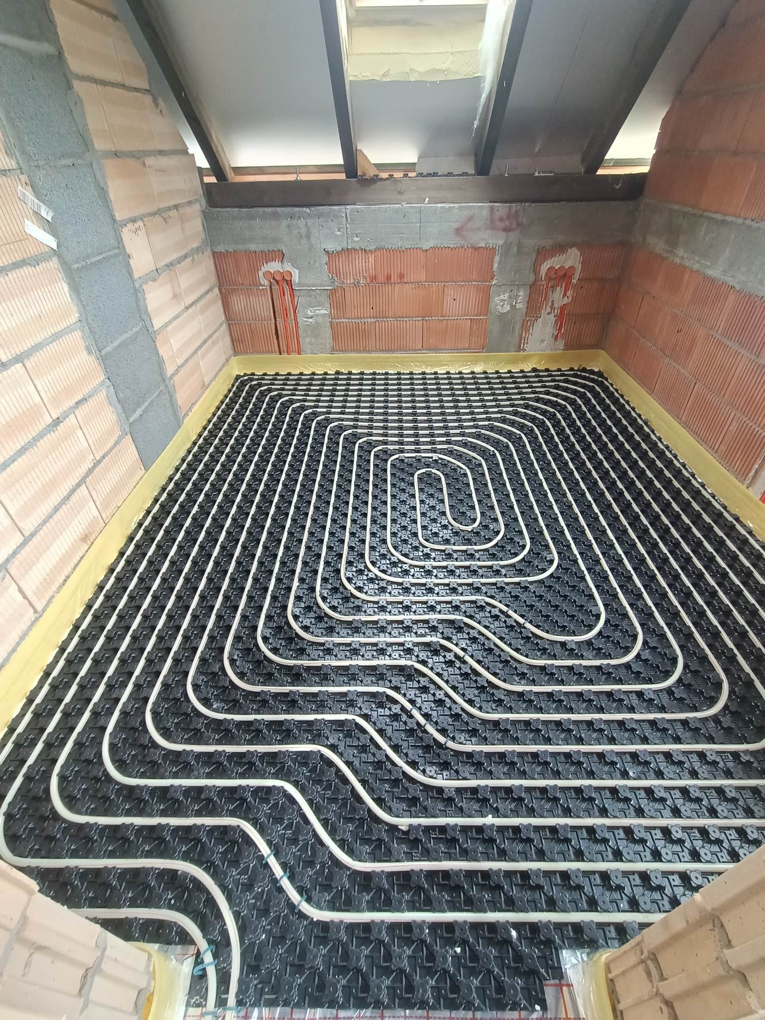 Rozłożona instalacja rur w systemie ogrzewania podłogowego na czarnej macie izolacyjnej, widoczne ceglane ściany w surowym stanie i elementy konstrukcji dachu.