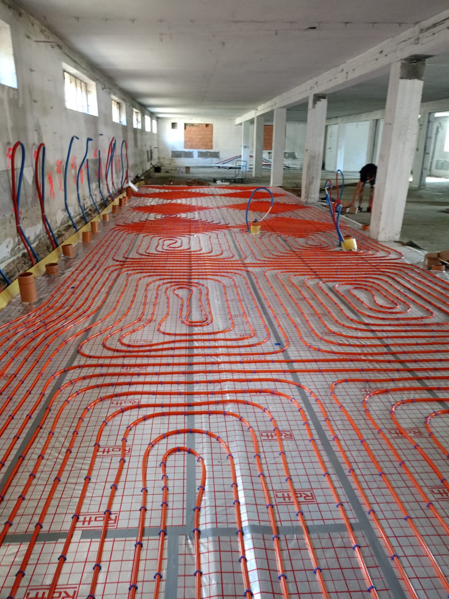 Instalacja rur grzewczych w systemie podłogowym na folii z siatką, widoczne rury doprowadzające w kolorach czerwonym i niebieskim, surowy stan budynku.