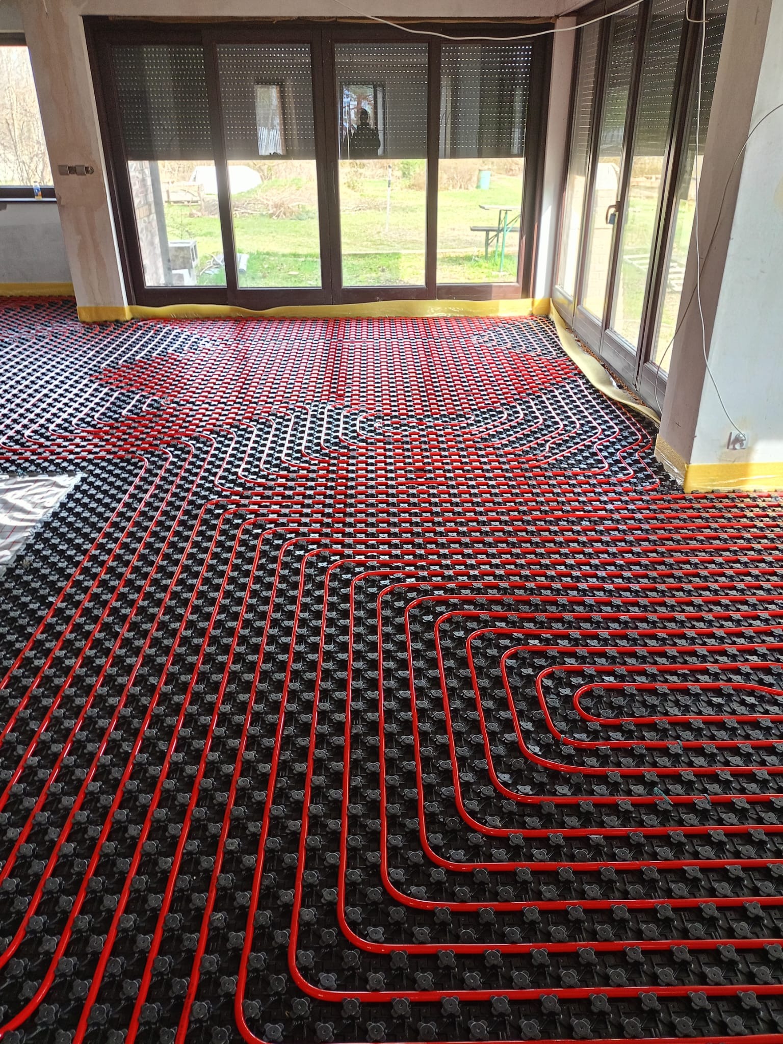 Czerwone rury ogrzewania podłogowego ułożone na czarnej macie w jasnym pomieszczeniu z dużymi oknami i żółtą folią ochronną przy ścianach.