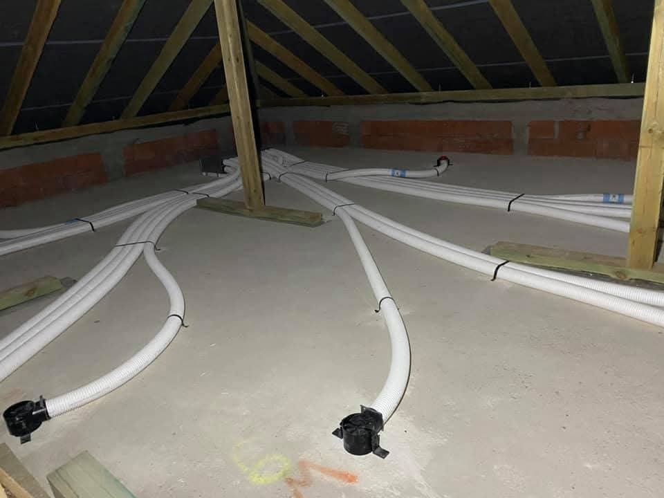 Białe, karbowane rury prowadzone po podłodze poddasza w Żarkach, zabezpieczone czarnymi opaskami zaciskowymi, widoczne elementy konstrukcji dachu i ceglanej ściany.