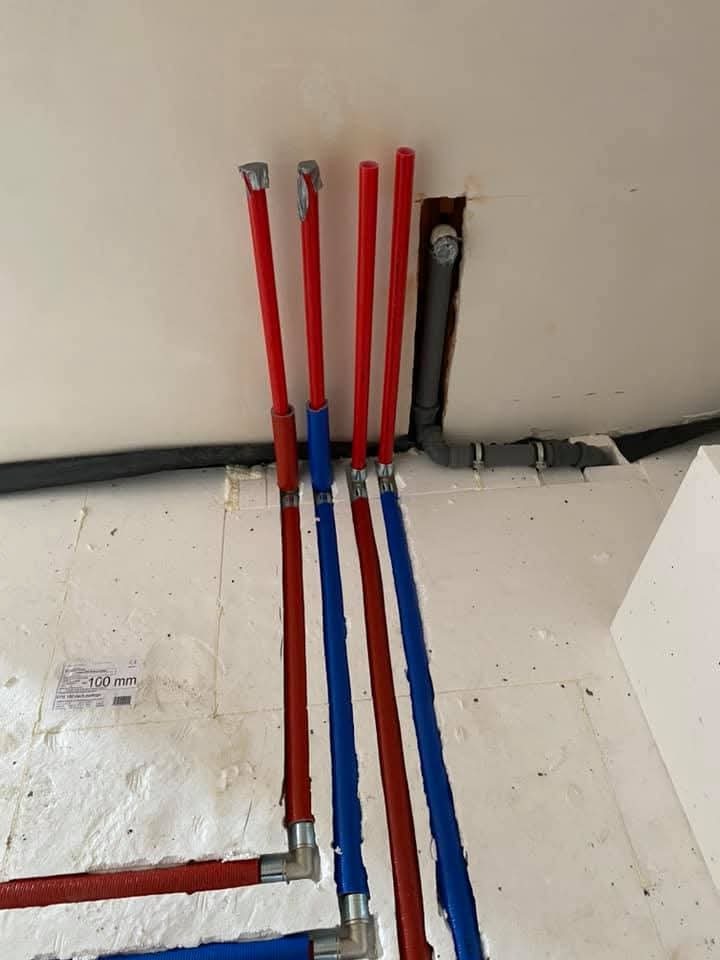 Instalacja rur PEX czerwonych i niebieskich w bruzdach w ścianie i podłodze, z widocznymi złączkami i fragmentem instalacji kanalizacyjnej w tle.