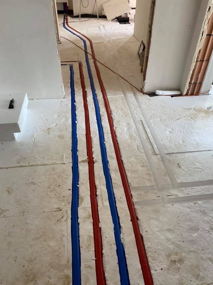 Instalacja rur grzewczych w izolacji podłogowej, widoczne rury w kolorze czerwonym i niebieskim, w trakcie montażu w nowym budownictwie.