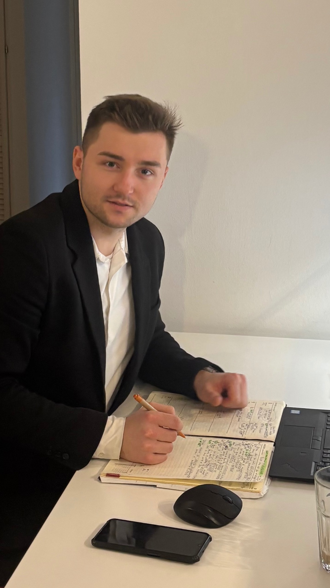 Młody doradca w garniturze, notuje w otwartym notatniku przy biurku z laptopem i telefonem. Przyjazny wyraz twarzy, jasne tło.