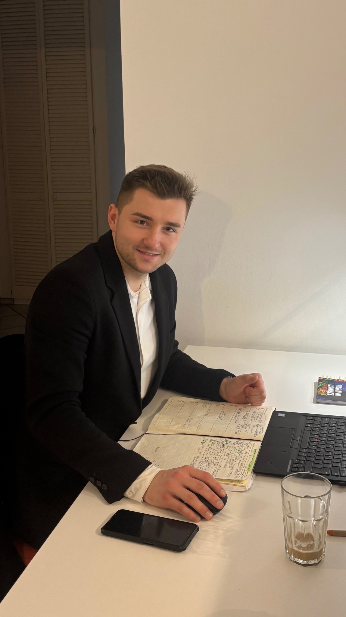 Doradca finansowy z Poznania uśmiecha się znad biurka z notatnikiem, laptopem i szklanką. Mężczyzna w garniturze, gotowy do analizy finansowej.