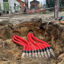 Wykop z ułożonymi czerwonymi rurami kablowymi w ziemi, widoczne oznaczenia na rurach, w tle torowisko tramwajowe i zabudowania miejskie.