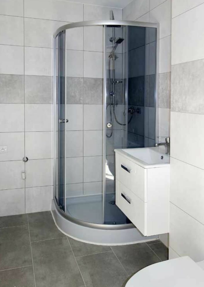 Nowoczesna łazienka z kabiną prysznicową narożną, białą szafką pod umywalkę z dwiema szufladami i szarymi płytkami na ścianach i podłodze.