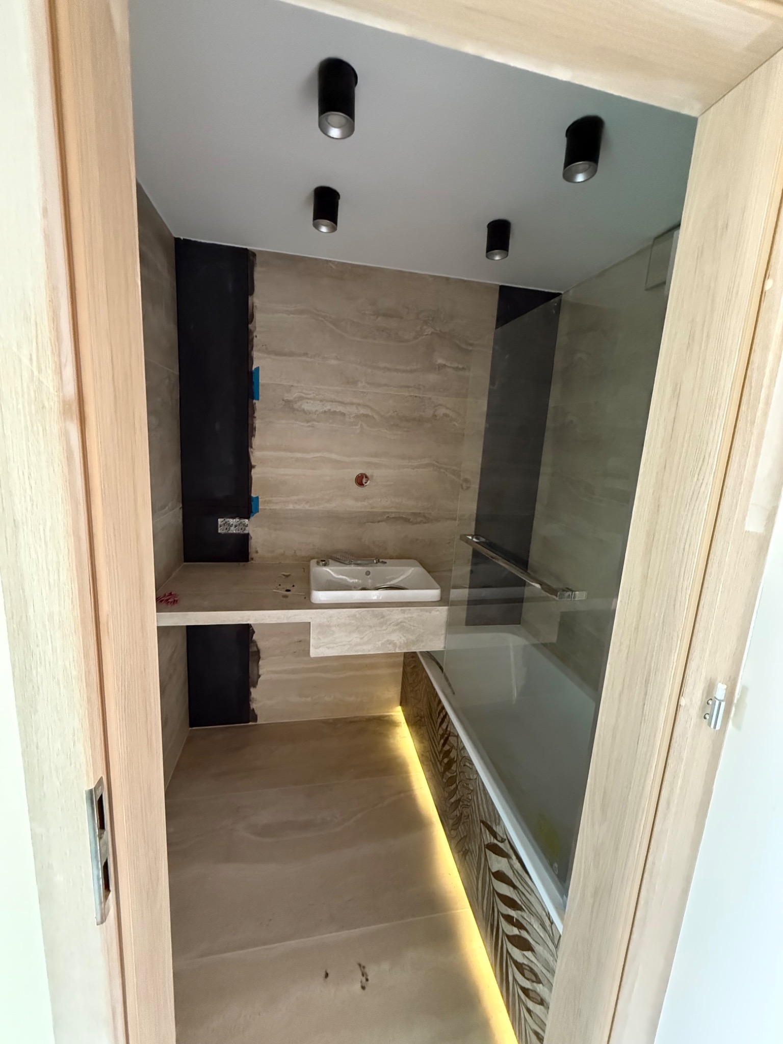 Nowoczesna łazienka w trakcie wykańczania: beżowe płytki, czarne akcenty, wanna z szybą, oświetlenie punktowe. Widok przez drewniane futryny drzwi.