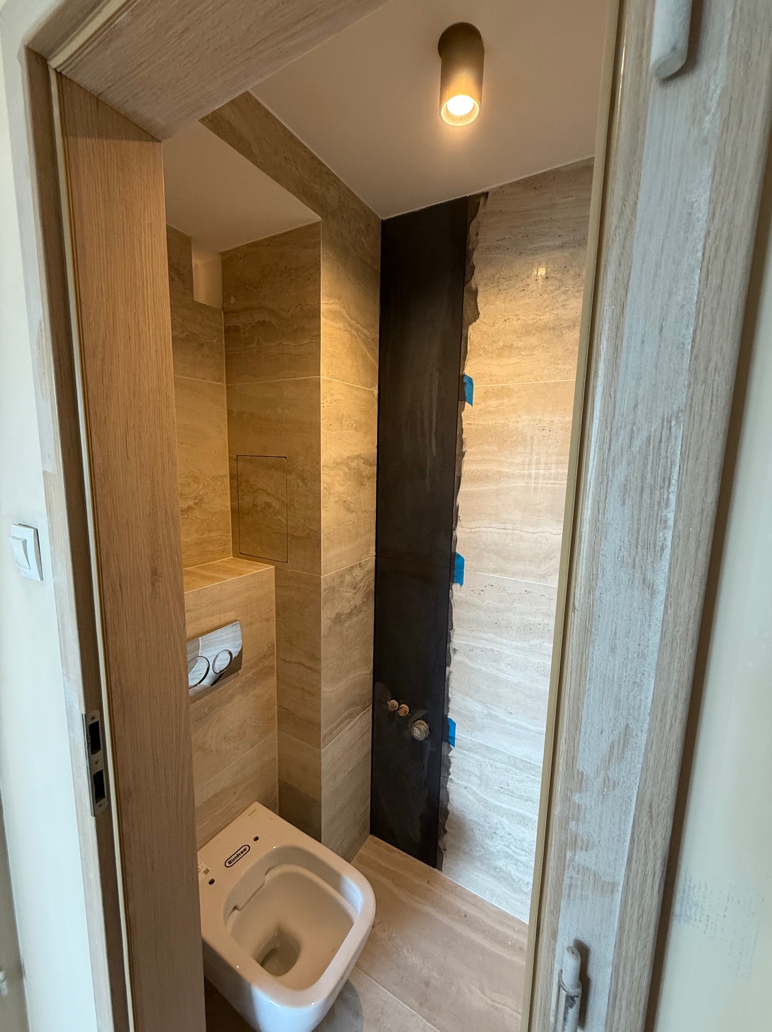 Nowoczesna łazienka w trakcie wykańczania, widoczne płytki imitujące drewno, częściowo zamontowana toaleta i fragment ściany przygotowany pod wykończenie.