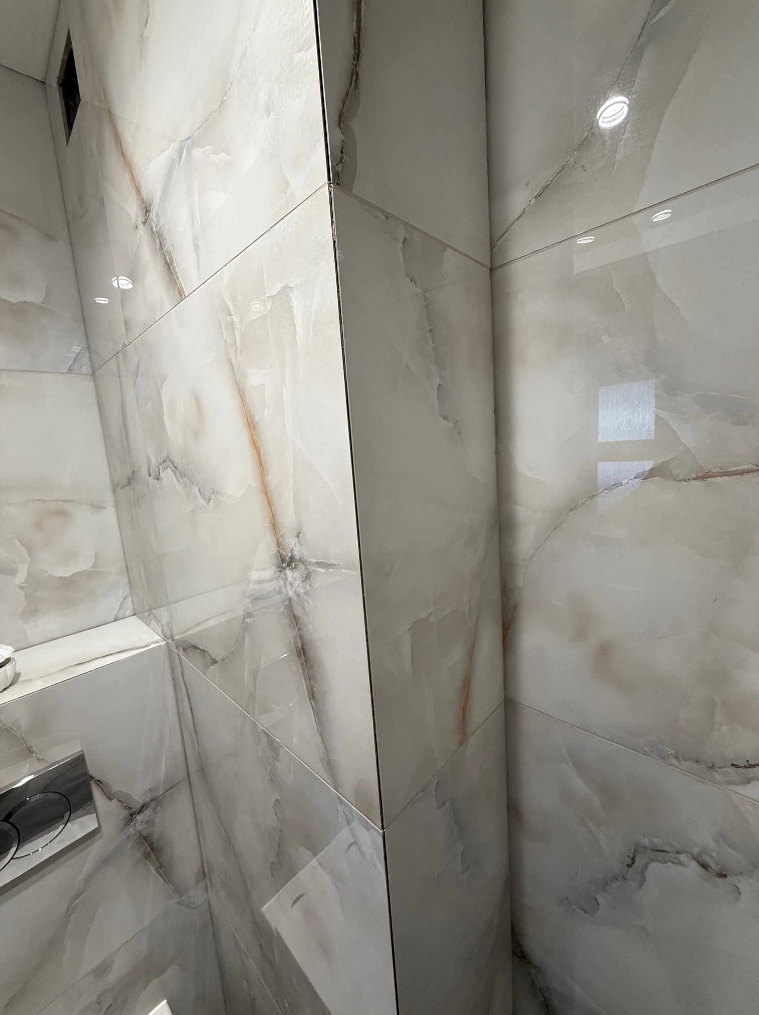 Wykończona łazienka z dużymi, białymi płytkami imitującymi marmur z delikatnymi, brązowymi żyłkami. Widoczny fragment toalety z chromowanym przyciskiem.