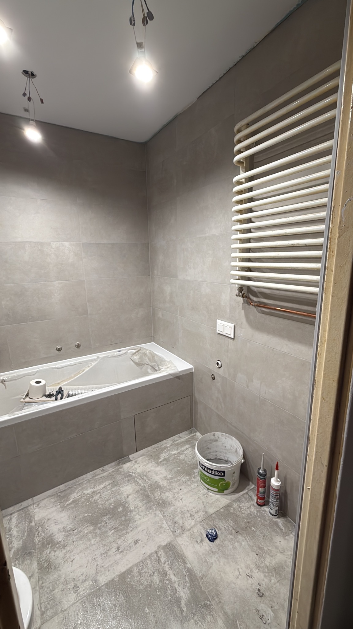 Łazienka w trakcie remontu: szare płytki, wanna zabezpieczona folią, grzejnik drabinkowy, wiadro farby Śnieżka i tuby silikonu na podłodze. Widoczne niedokończone oświetlenie.