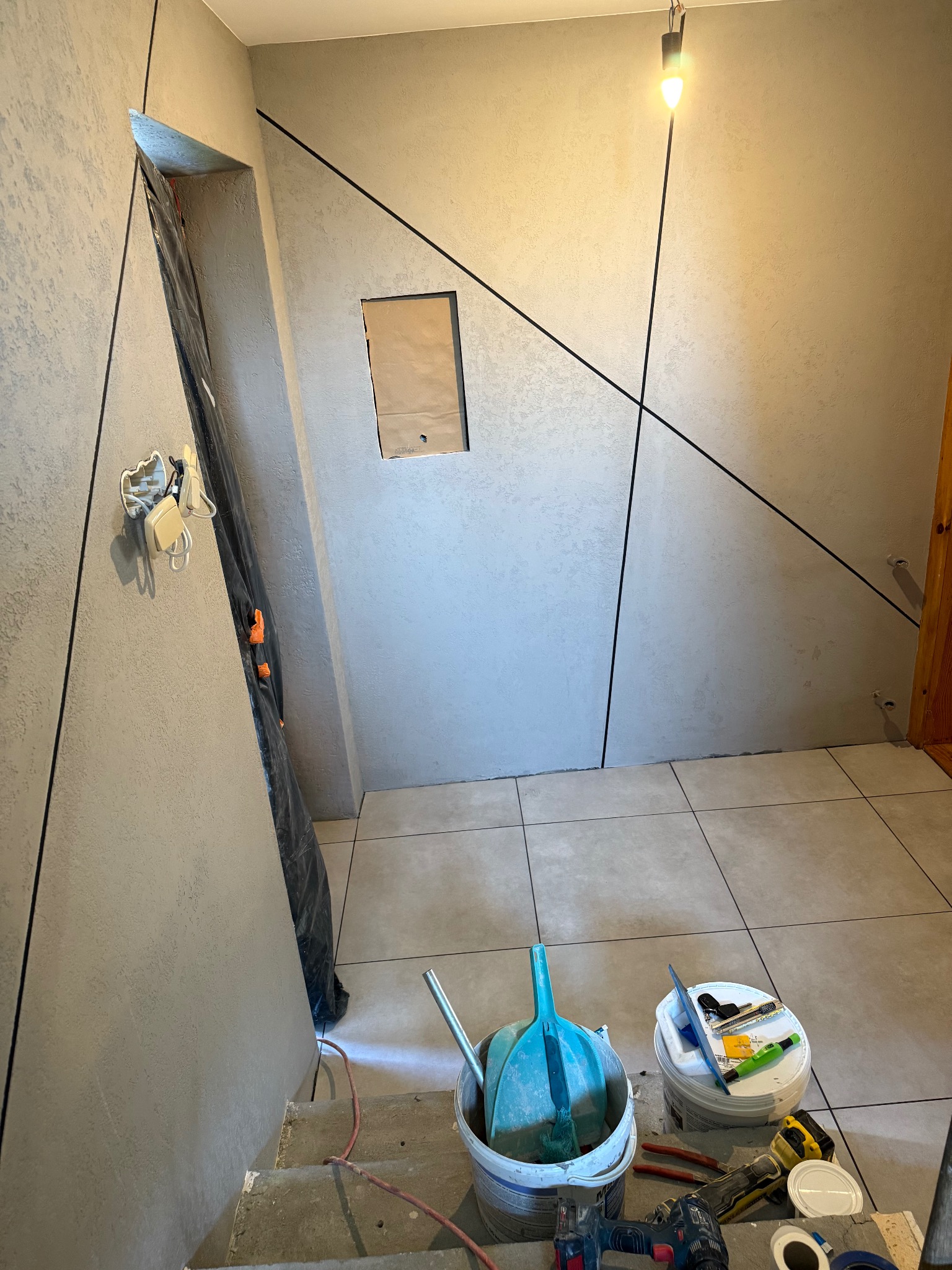 Wnętrze w trakcie wykończenia: szare ściany z dekoracyjnymi czarnymi liniami, częściowo położona podłoga z płytek, narzędzia i akcesoria malarskie na schodach.