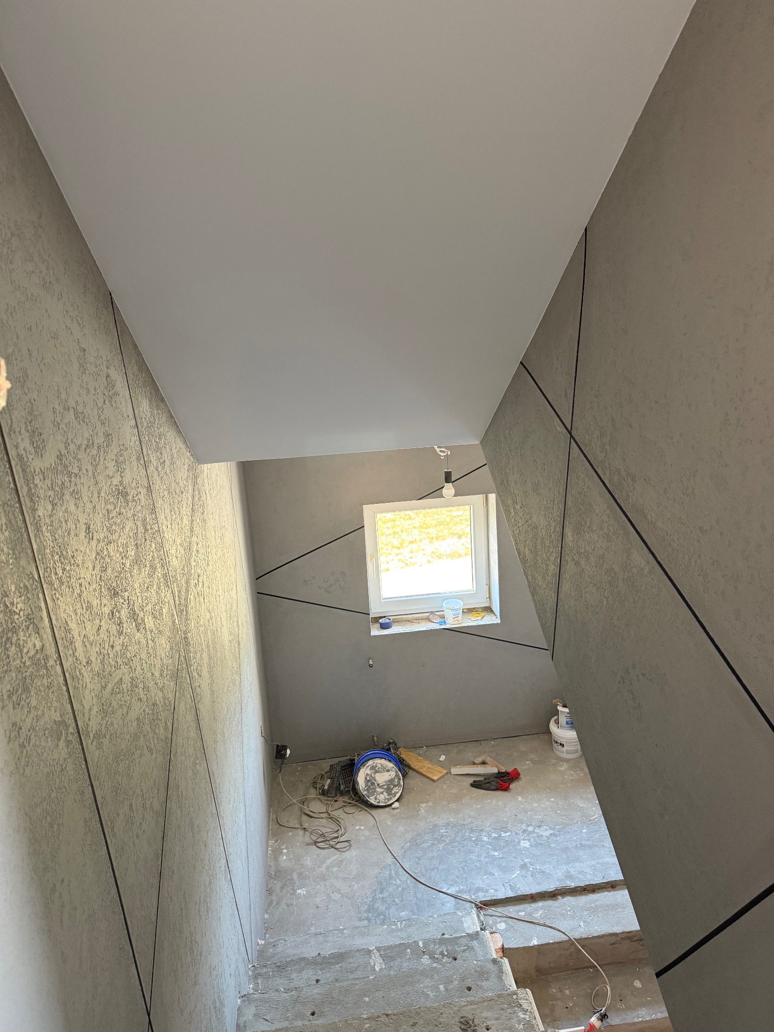 Klatka schodowa w trakcie wykończenia: szare ściany z dekoracyjnymi czarnymi liniami, sufit pomalowany na biało, narzędzia budowlane na podłodze, okno w tle.