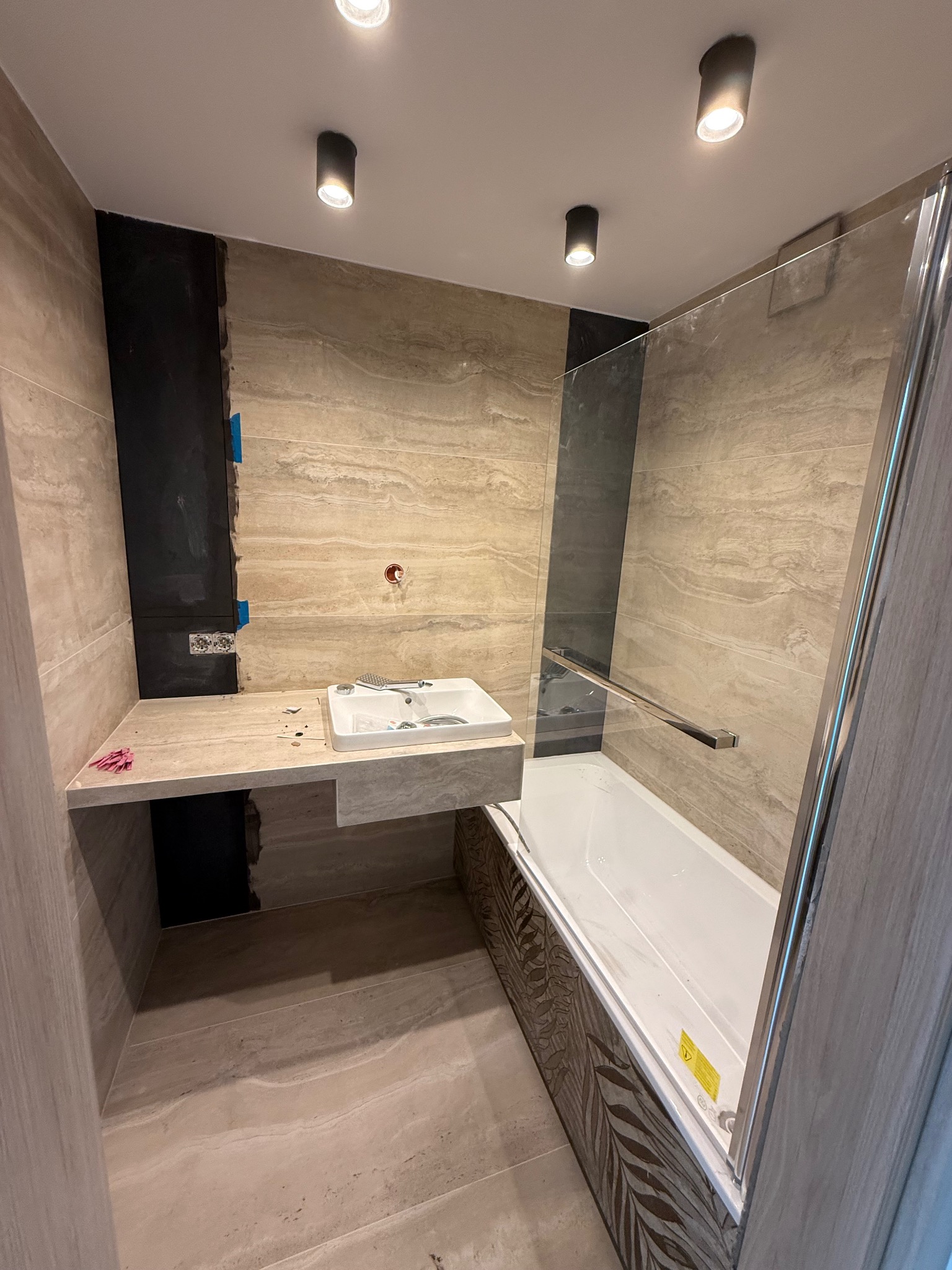 Wnętrze łazienki w trakcie wykańczania: zamontowana umywalka nablatowa, częściowo położone płytki, kabina nawannowa ze srebrnymi profilami.