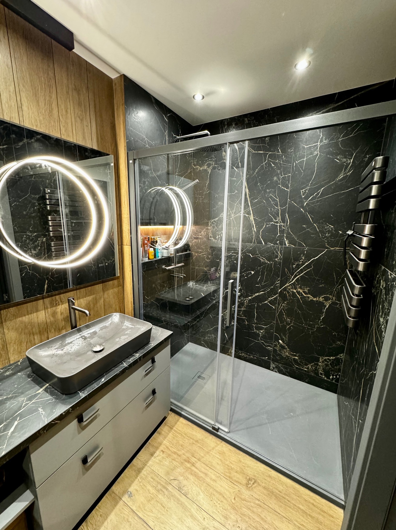 Nowoczesna łazienka z czarnymi marmurowymi płytkami, szarą umywalką nablatową i podświetlanym lustrem w okrągłej ramie, widoczny grzejnik drabinkowy i kabina prysznicowa.