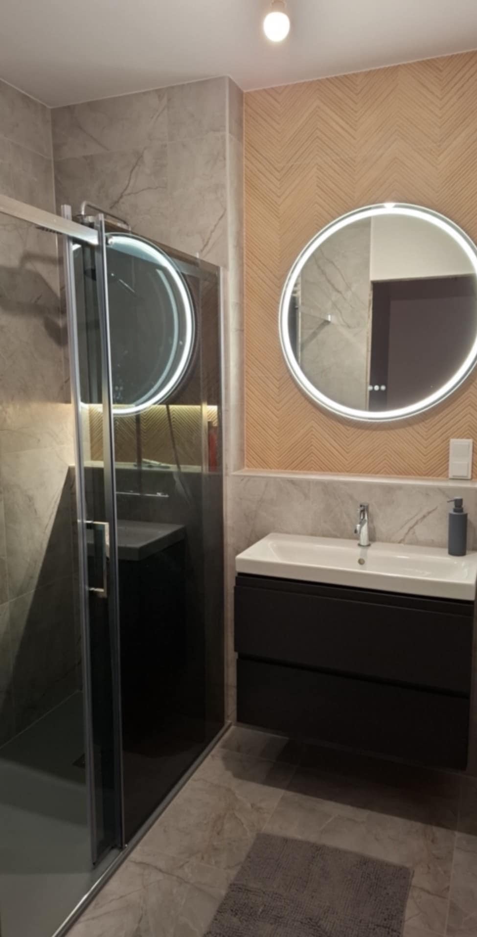 Nowoczesna łazienka z kabiną prysznicową z przesuwnymi drzwiami, okrągłym lustrem z podświetleniem LED, umywalką nablatową z szafką w kolorze antracytowym oraz ścianą z dekoracyjnym wzorem jodełki.