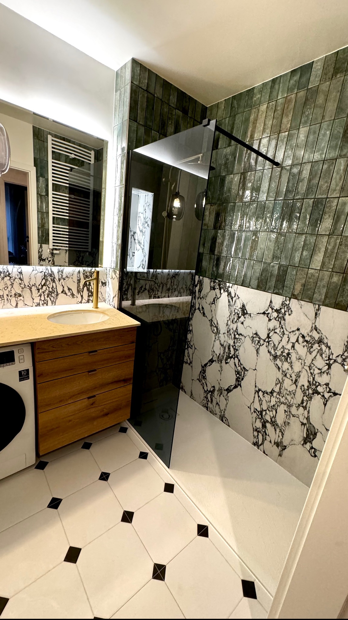 Nowoczesna łazienka z zielonymi, podłużnymi płytkami ułożonymi pod kątem, kabiną prysznicową typu walk-in z czarnymi elementami, umywalką nablatową na drewnianej szafce z pralką obok i podłogą...