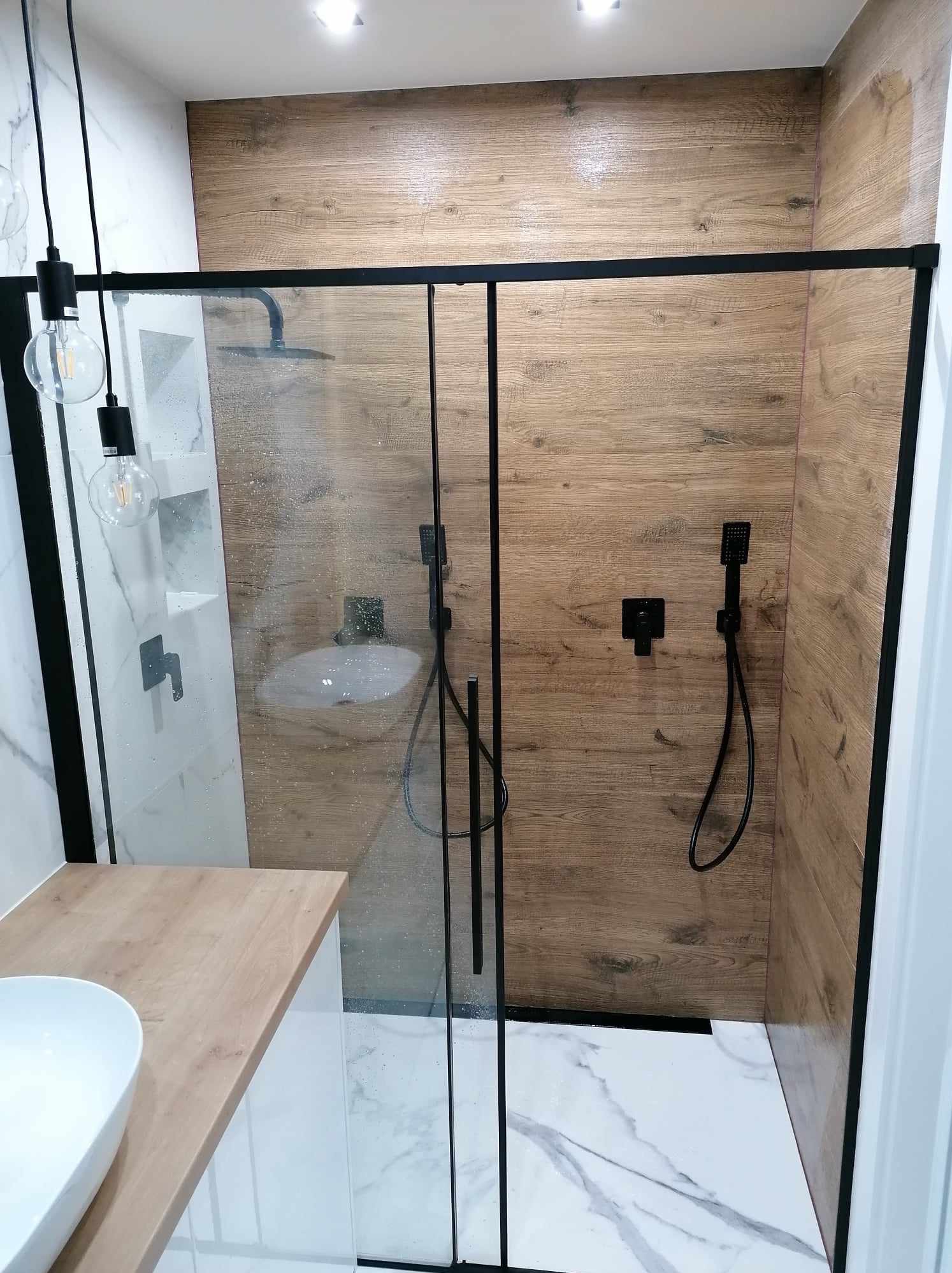 Nowoczesna kabina prysznicowa z czarnymi ramami i armaturą, drewnopodobne płytki ścienne, marmurowa podłoga i umywalka nablatowa z drewnianym blatem.