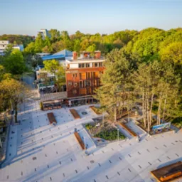 Widok z lotu ptaka na odnowiony plac miejski z budynkiem apartamentowym w tle, otoczony zielenią drzew, z drewnianymi ławkami i nowoczesnym oświetleniem.