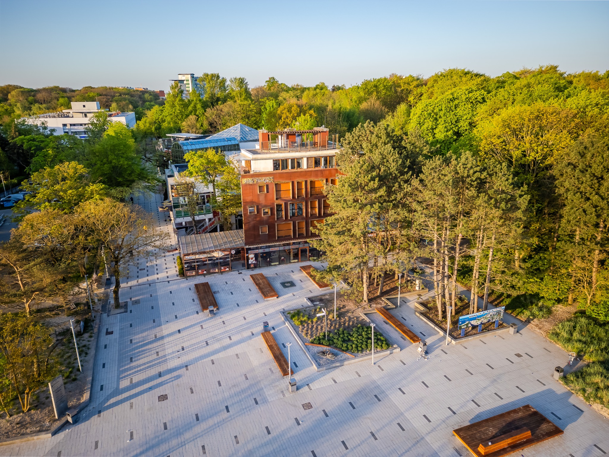 Widok z lotu ptaka na odnowiony plac miejski z budynkiem apartamentowym w tle, otoczony zielenią drzew, z drewnianymi ławkami i nowoczesnym oświetleniem.