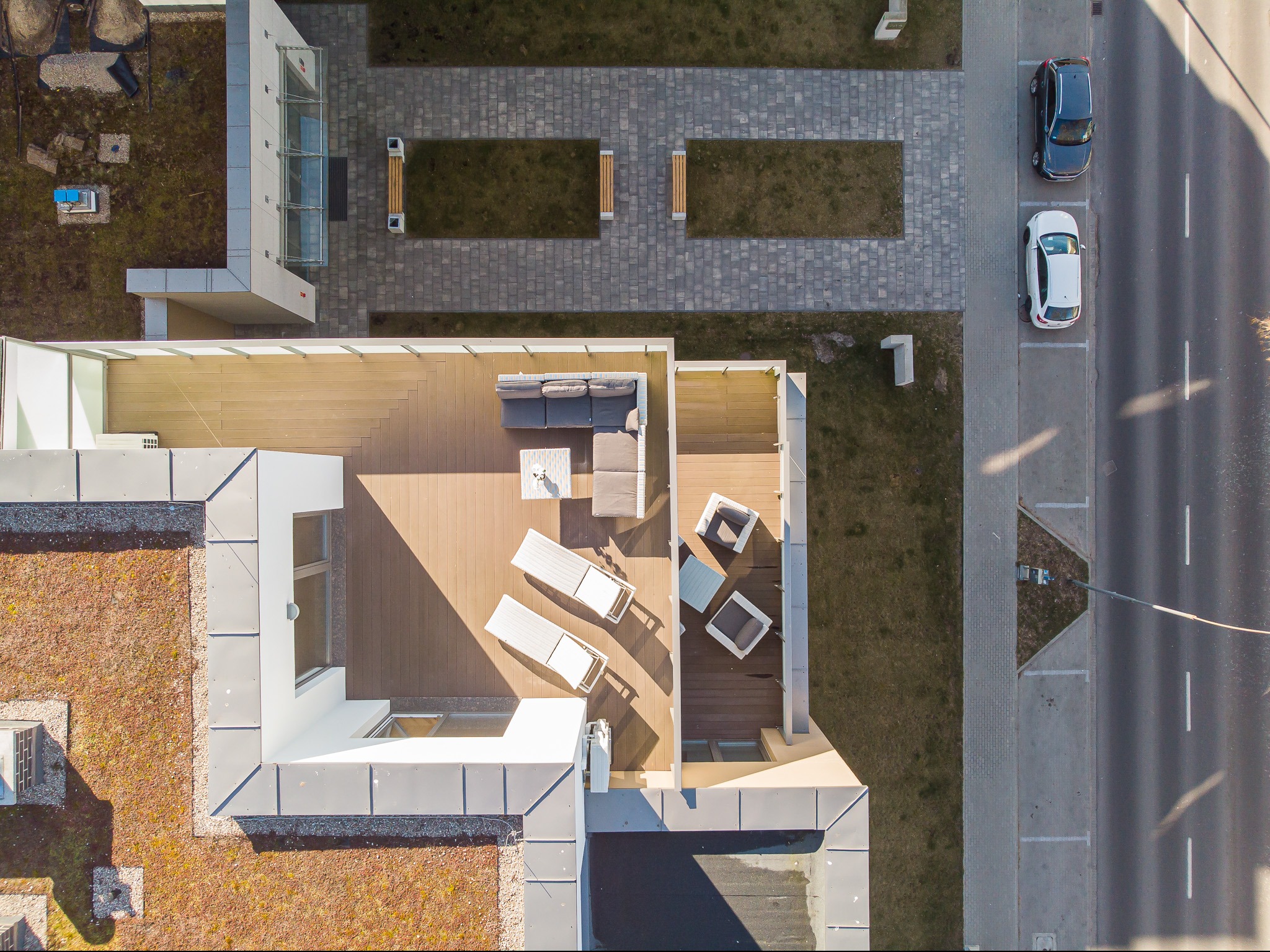 Widok z lotu ptaka na taras z drewnianą podłogą, białymi leżakami, szarą sofą i zielony dach budynku obok ulicy z zaparkowanymi samochodami.