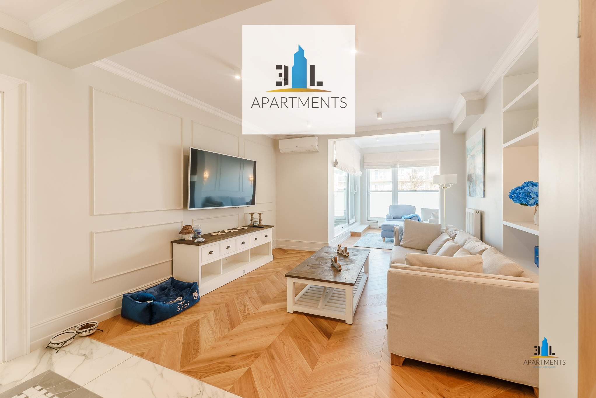 Jasny salon w apartamencie z parkietem w jodełkę, beżową sofą, telewizorem i niebieskim legowiskiem dla psa obok szafki RTV.