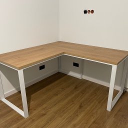 R.A.R.Z. - Nowoczesne biurko narożne z blatem imitującym dąb i białymi metalowymi nogami w minimalistycznym wnętrzu, widoczne gniazdka elektryczne bez osłony na ścianie.