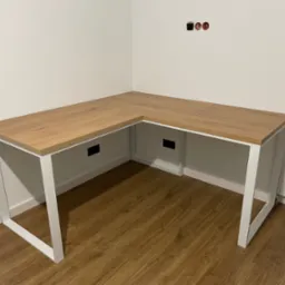 Nowoczesne biurko narożne z blatem imitującym dąb i białymi metalowymi nogami w minimalistycznym wnętrzu, widoczne gniazdka elektryczne bez osłony na ścianie.