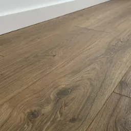 Ułożone panele podłogowe w odcieniach brązu, z widoczną strukturą drewna i białymi listwami przypodłogowymi przy ścianie.