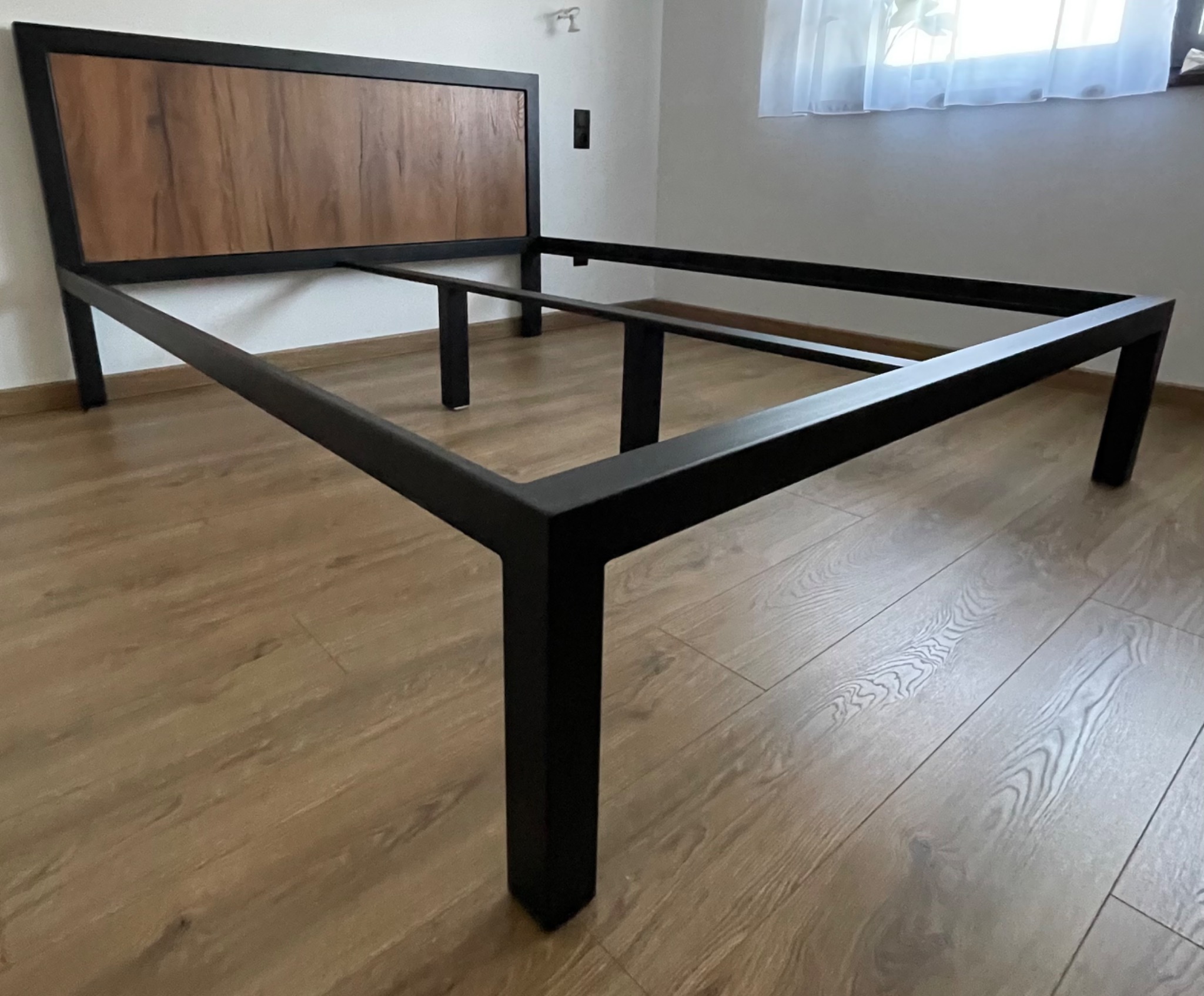 Rama łóżka z czarnego metalu z drewnianym zagłówkiem, ustawiona na podłodze z paneli imitujących drewno, w pokoju z białymi ścianami i oknem z firanką.