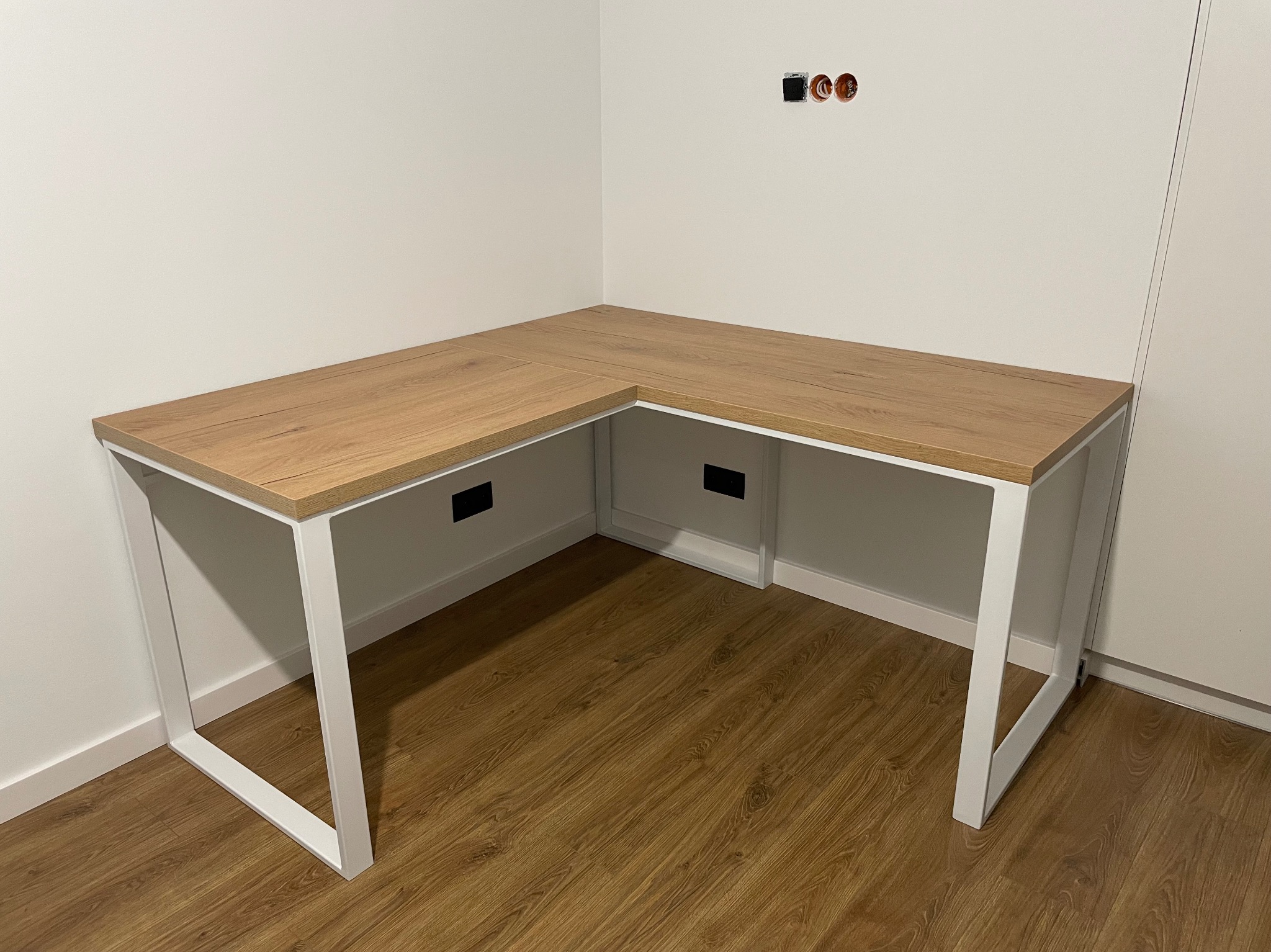 Nowoczesne biurko narożne z blatem imitującym dąb i białymi metalowymi nogami w minimalistycznym wnętrzu, widoczne gniazdka elektryczne bez osłony na ścianie.