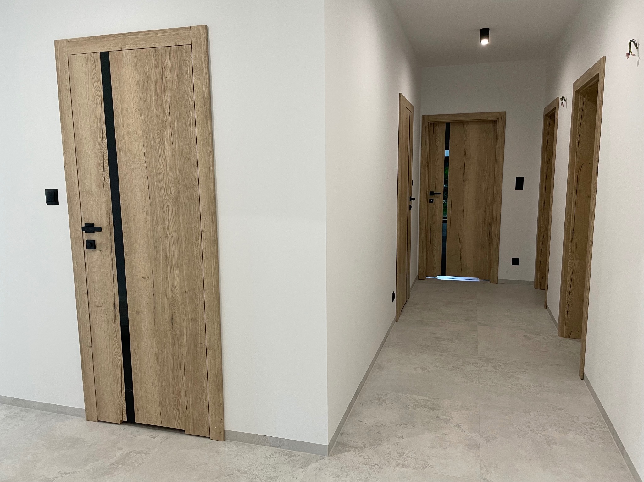 Wnętrze korytarza z trzema drewnianymi drzwiami z czarnymi wstawkami, szare płytki na podłodze, białe ściany i minimalistyczne oświetlenie sufitowe.