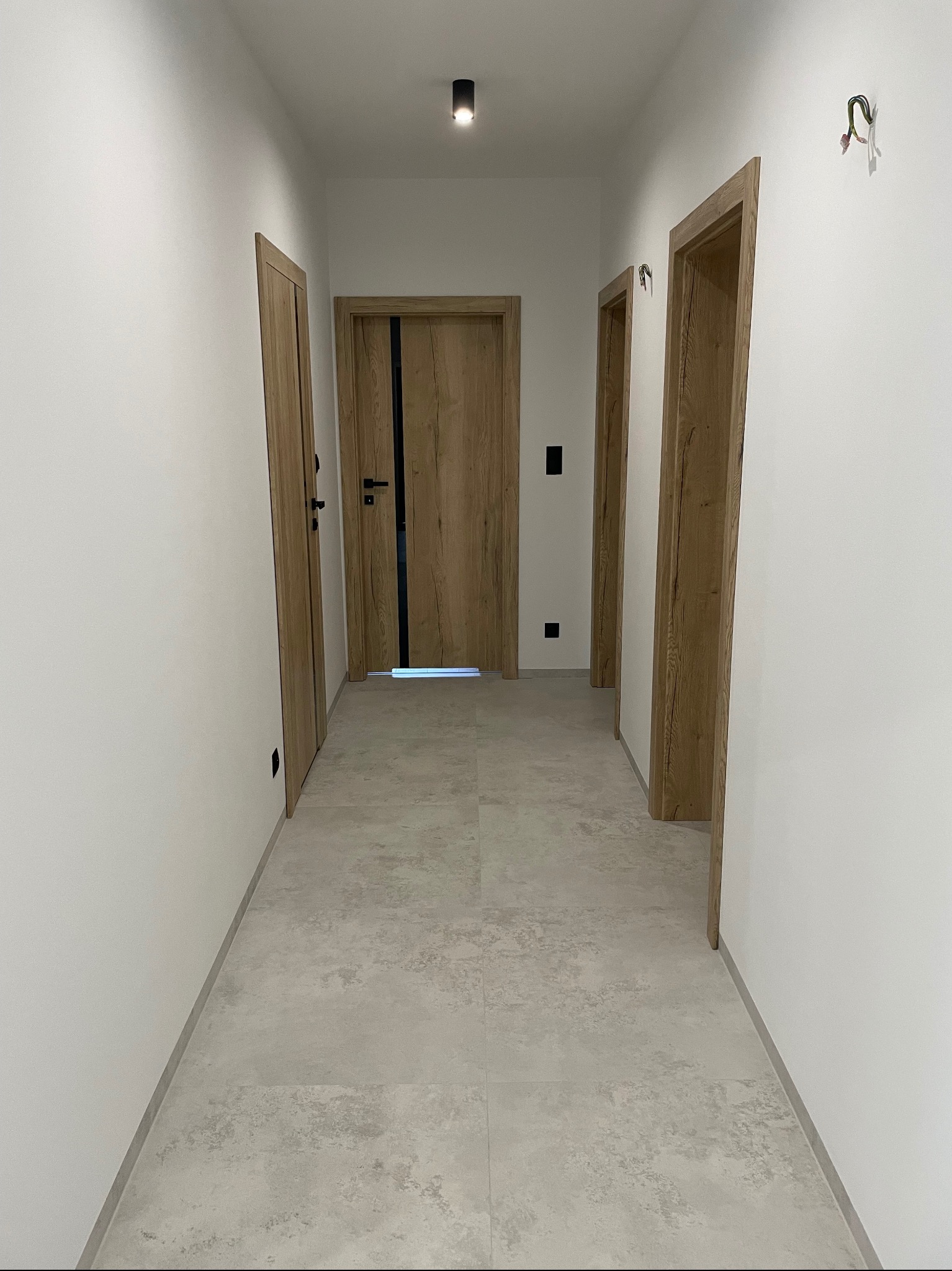 Jasny korytarz z trzema drewnianymi drzwiami, wykończony szarą podłogą z płytek imitujących kamień, białe ściany z wystającymi przewodami elektrycznymi i czarnym oświetleniem punktowym na suficie.