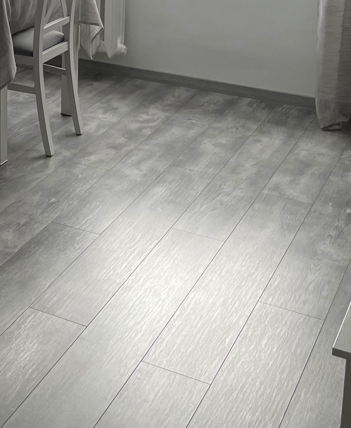 Jasnoszara podłoga z paneli ułożonych wzdłuż, widok z perspektywy, fragment krzesła z jasnego drewna i zasłony w tle.