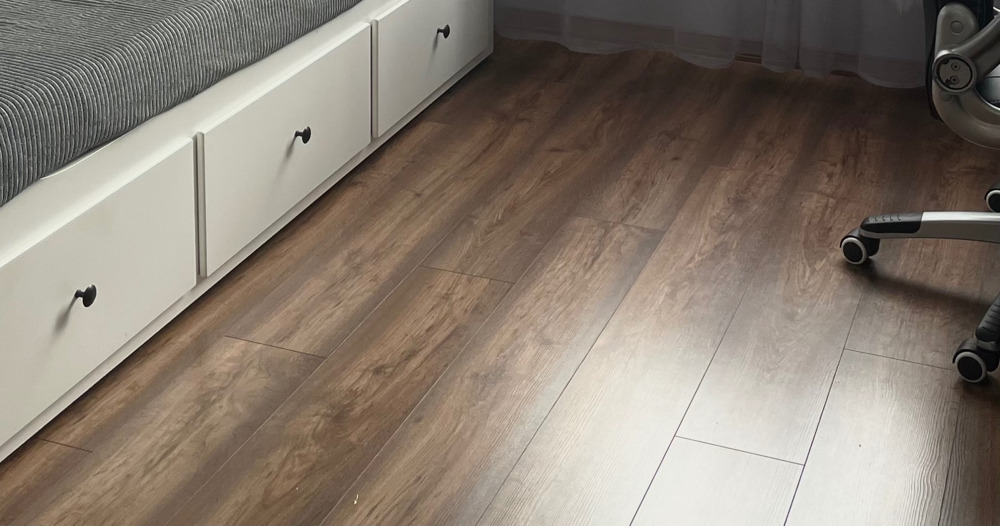 Ułożone panele podłogowe w odcieniach brązu, widoczne fragmenty białej zabudowy z szufladami pod łóżkiem oraz krzesło biurowe na kółkach.